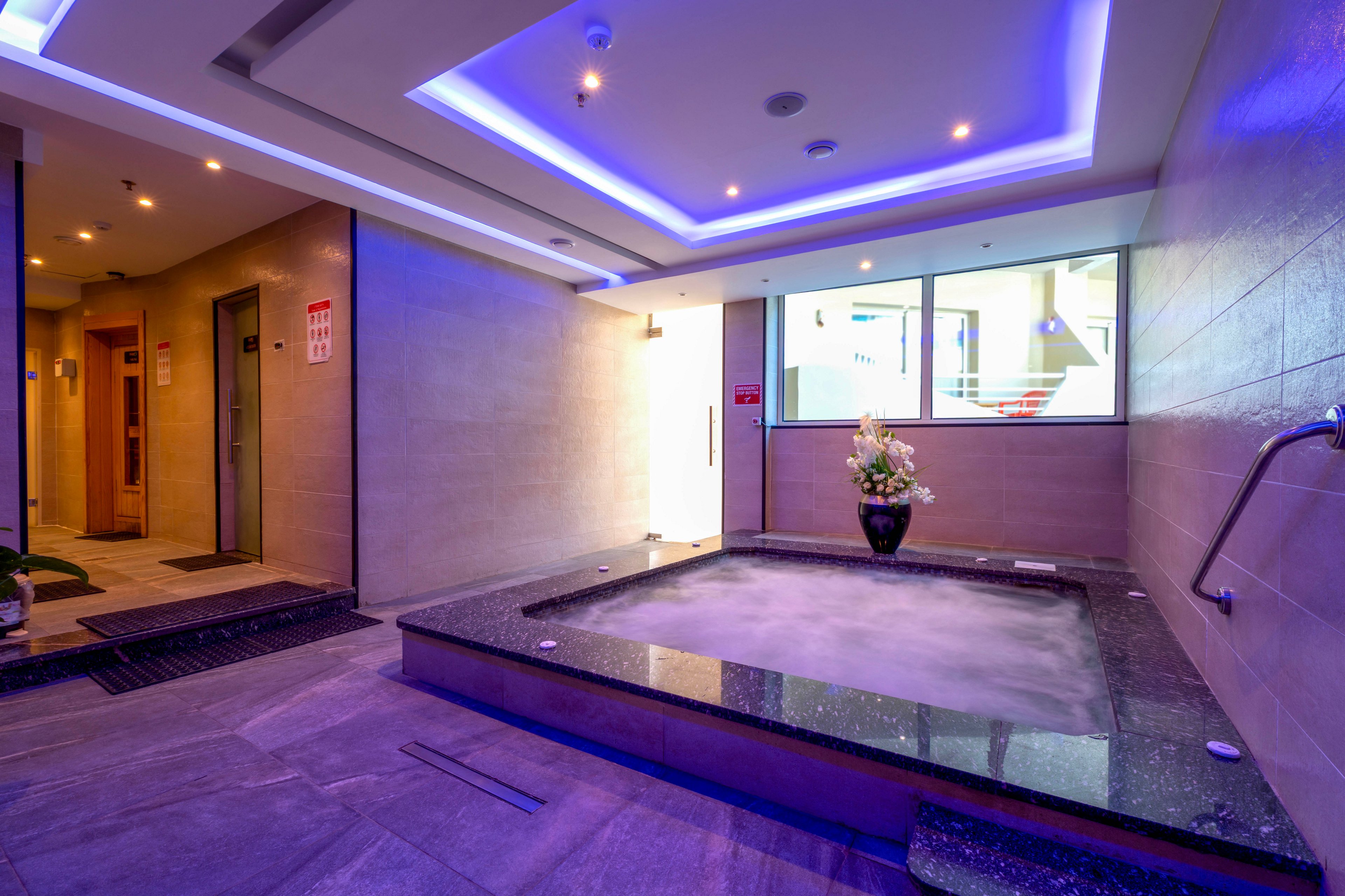 A contemporary indoor spa features a rectangular hot tub with bubbling water, surrounded by sleek stone tiles. The setting is enhanced by recessed blue ceiling lights and a large window providing natural light. A decorative vase with flowers sits at the corner of the tub, adding a touch of elegance. The atmosphere is tranquil and inviting, ideal for relaxation.