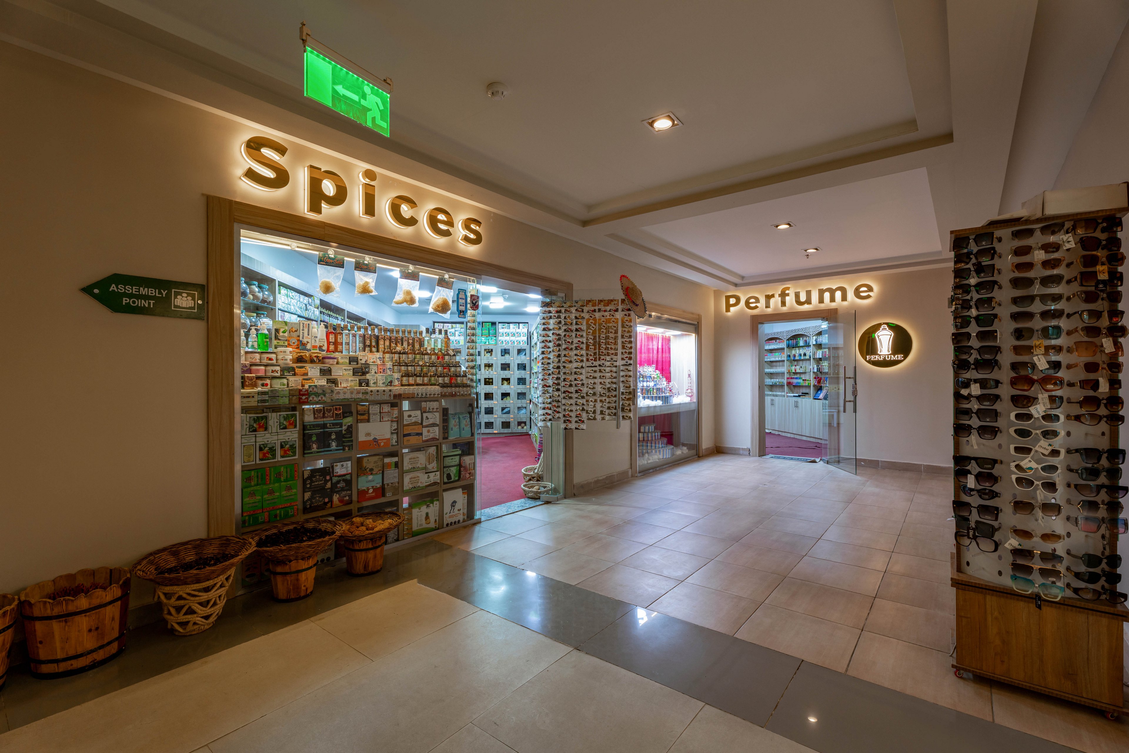 This image shows a well-lit indoor shopping corridor featuring two adjacent stores labeled 'Spices' and 'Perfume.' The spices shop displays shelves filled with jars and containers, while the perfume shop has a modern, inviting entrance. Sunglasses are displayed on a rotating rack in the foreground, and a green exit sign is visible above. The overall atmosphere is clean and organized, with neutral tones and clear signage.