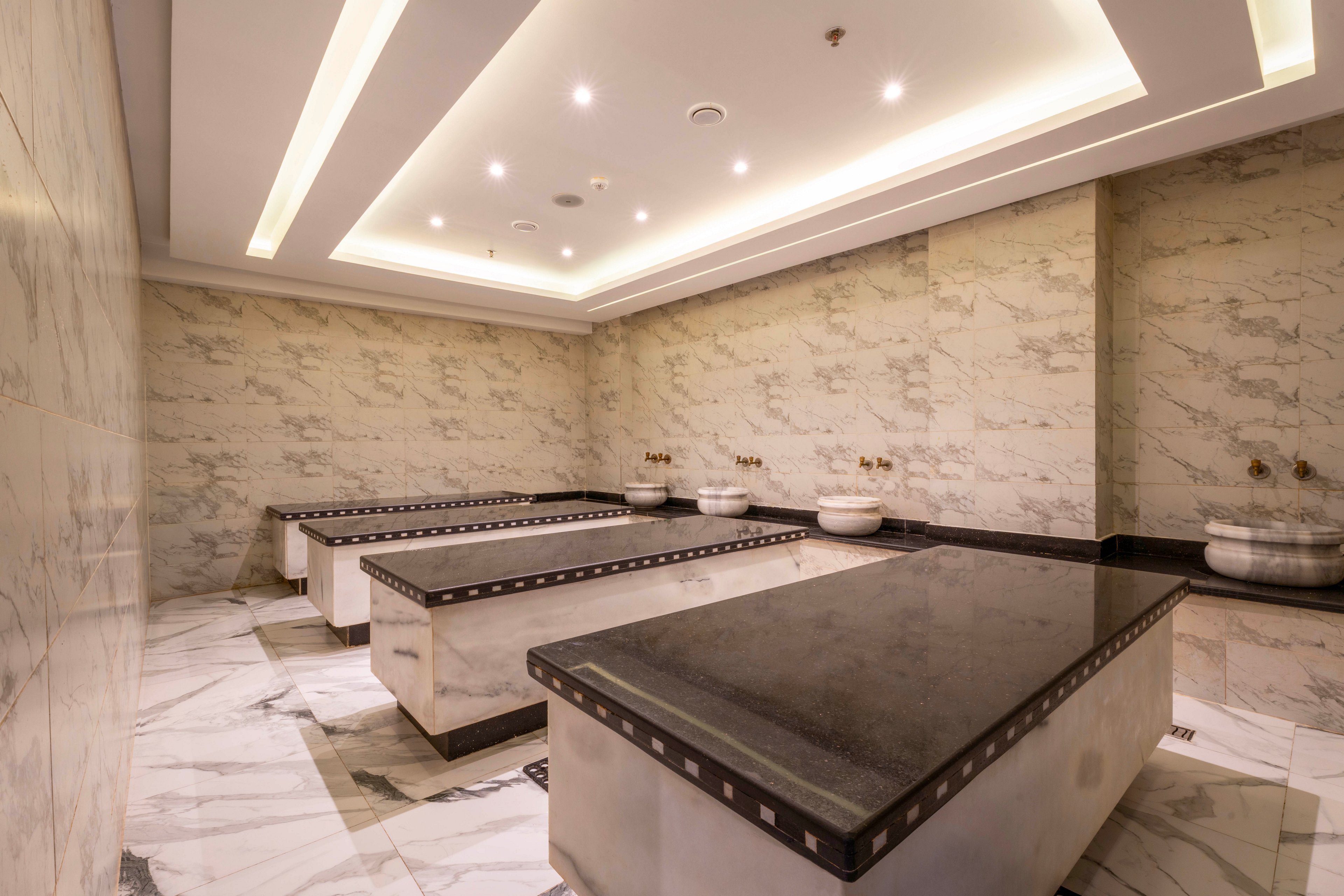 This image shows a spacious Turkish bath, also known as a hammam, featuring multiple large marble massage tables. The setting is clean and modern, with polished marble walls and floors, recessed ceiling lighting, and traditional stone basins along the wall. The overall palette is neutral with white and gray tones, creating a serene and luxurious atmosphere. No people or visible text are present in the scene.