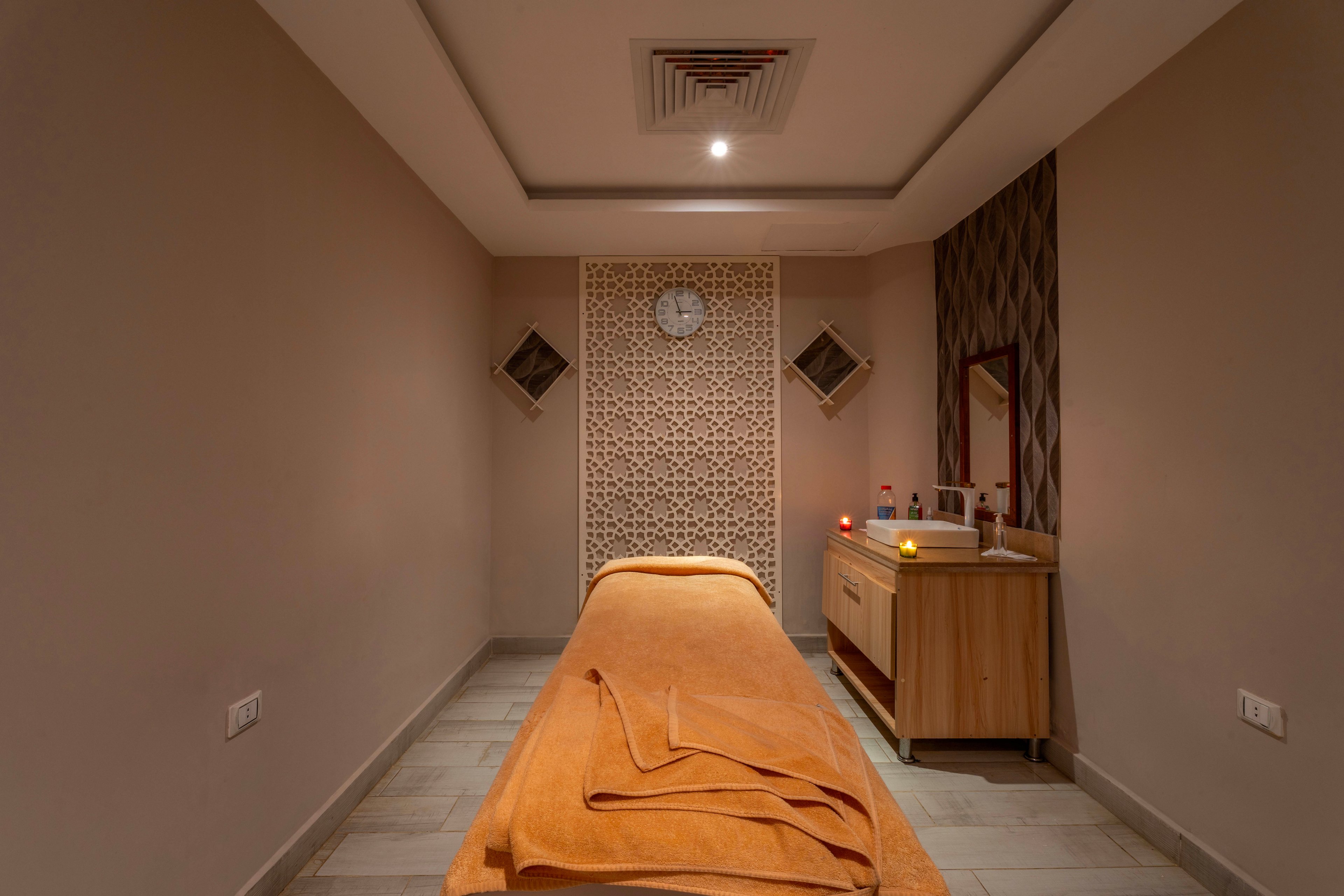 A neatly arranged massage table covered with orange towels is the focal point of this modern spa treatment room. The setting features soft, warm lighting, a decorative wall panel, and a small vanity with candles and amenities. A round wall clock is visible above the table, adding a functional touch to the serene environment. The overall mood is calm and inviting, ideal for relaxation and wellness treatments.