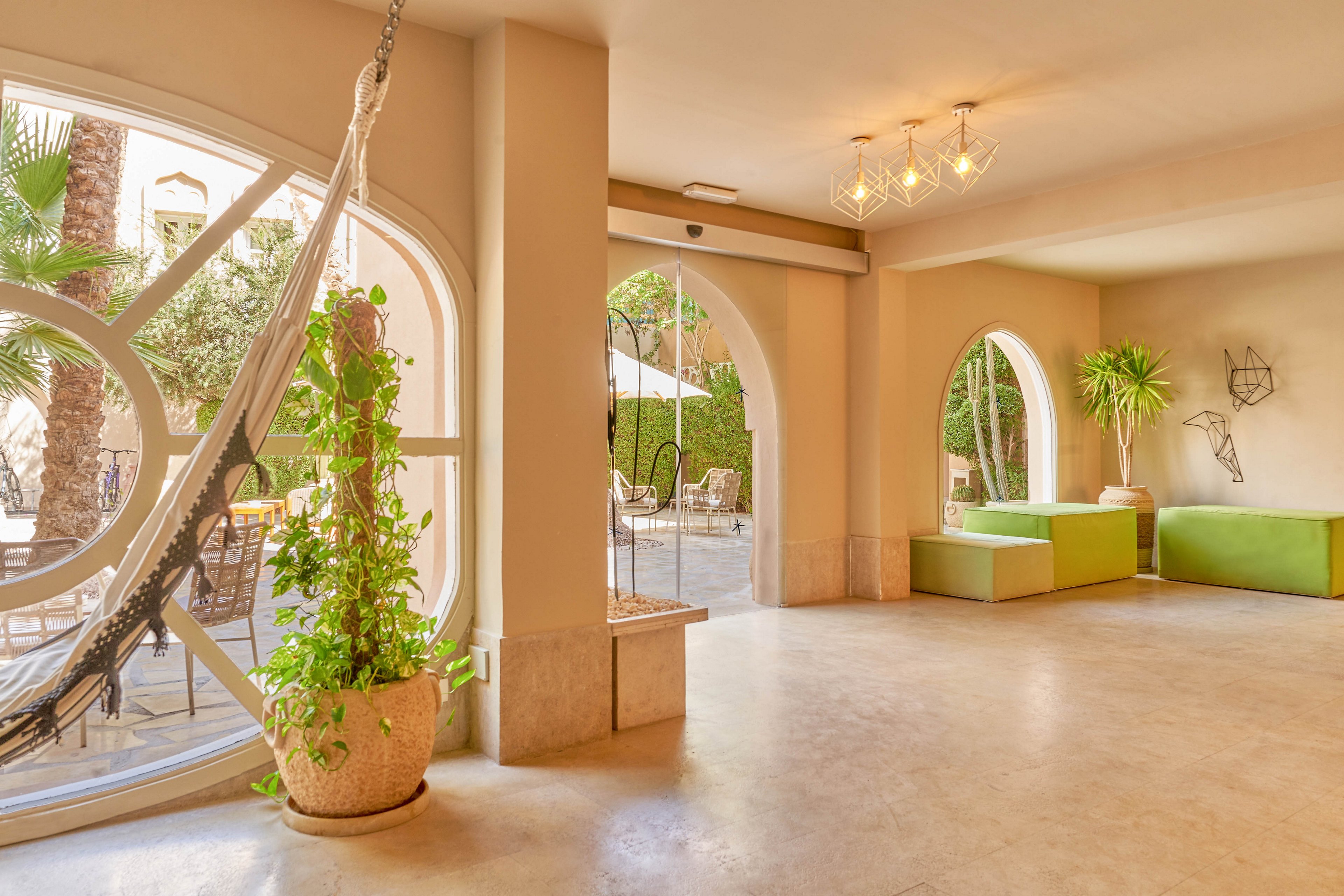 A spacious, sunlit lounge features a hammock suspended near a large circular window with lush green plants. The room is decorated in neutral tones with pops of green from minimalist seating and potted plants. Large arched windows offer views of an outdoor patio with tables and umbrellas. The overall mood is relaxed and inviting, with contemporary lighting fixtures and clean architectural lines.