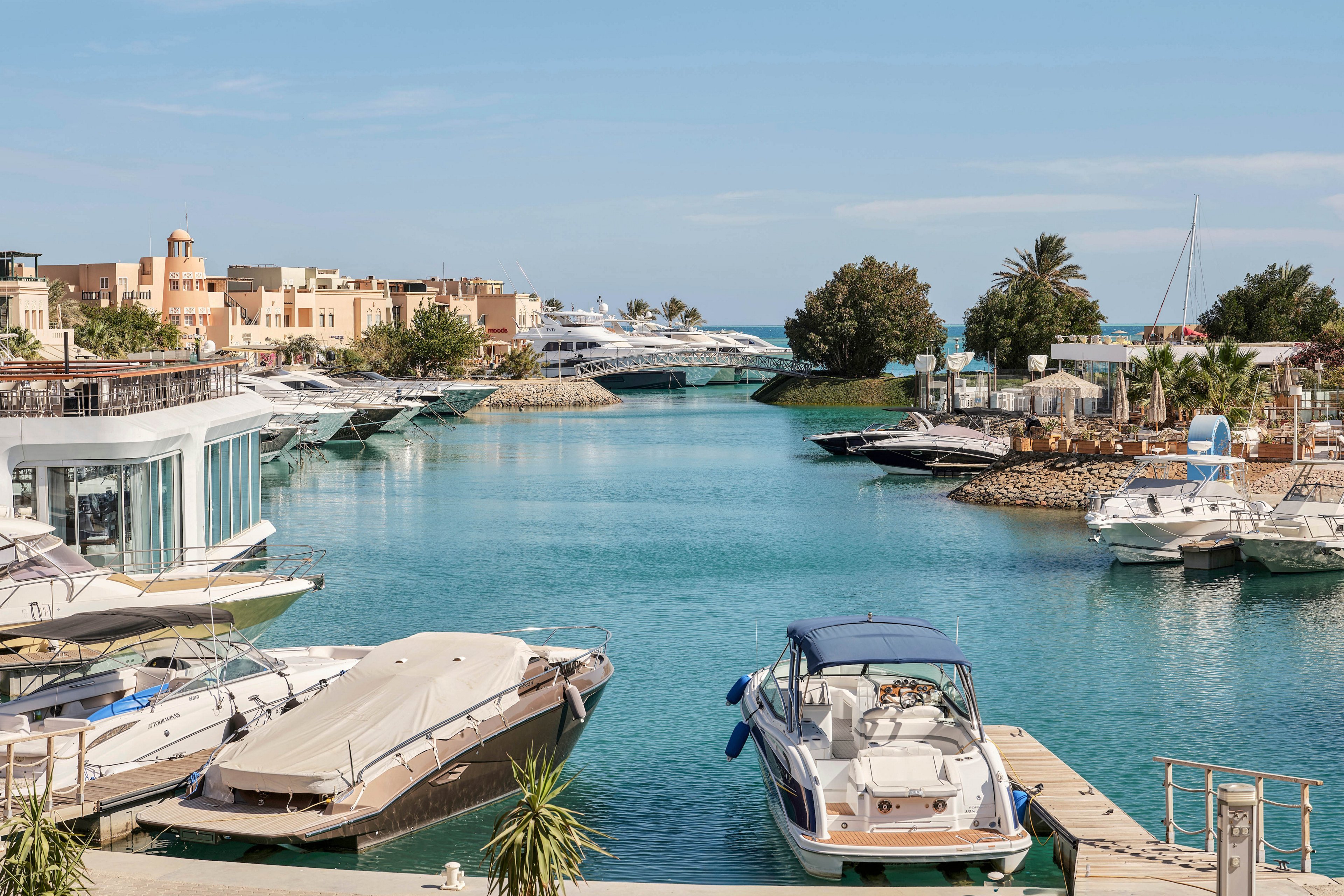 Several luxury yachts and boats are docked along a calm marina with turquoise water. The setting features modern waterfront buildings, palm trees, and a clear blue sky, creating a relaxed and upscale atmosphere. The scene is tranquil, with no visible people, and the architecture suggests a resort or exclusive residential area. No visible text or numbers are present in the image.