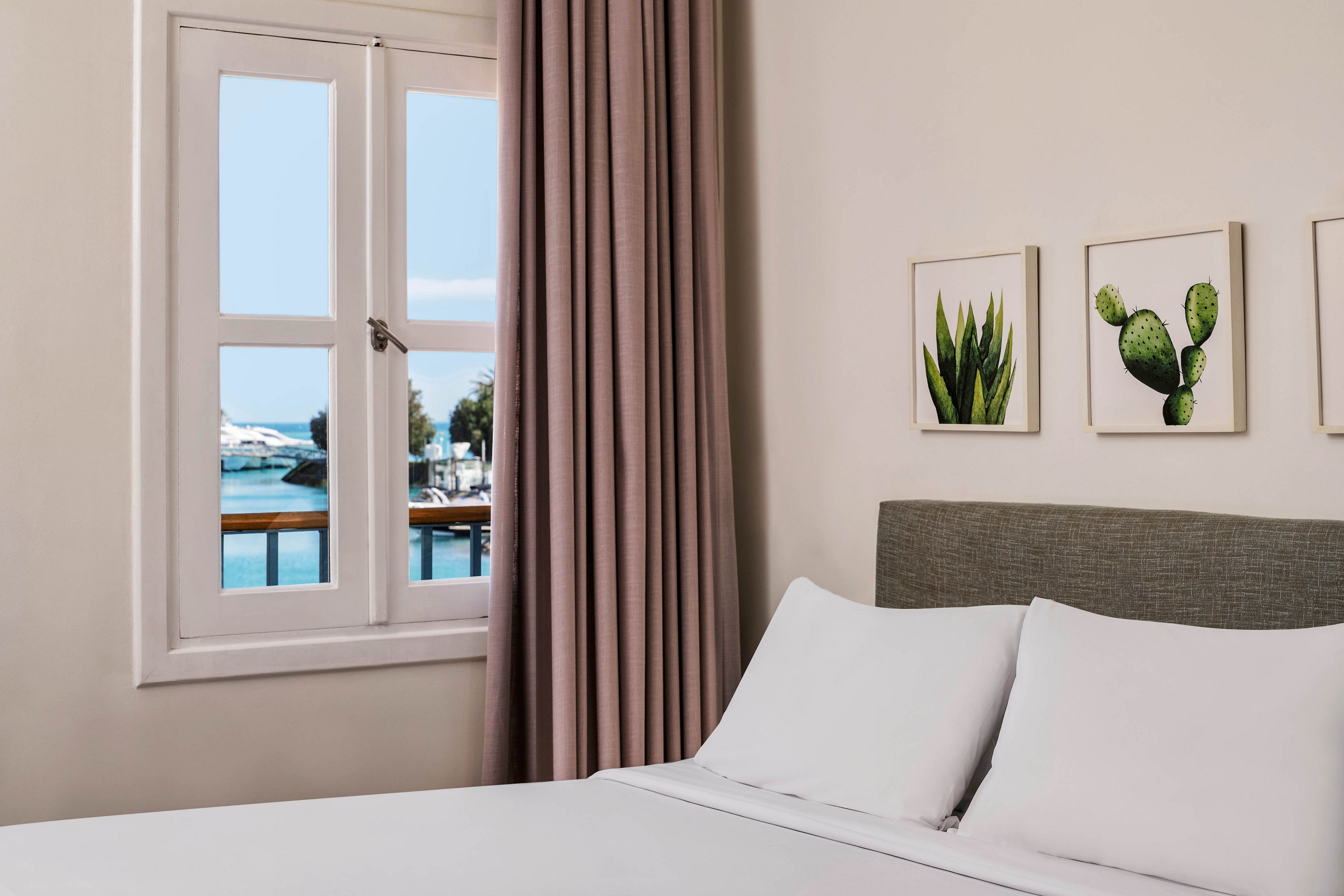 A neatly made bed with white linens sits beside a window overlooking a serene waterfront scene. The room features minimalist decor, including two framed botanical prints above the headboard and soft taupe curtains. Natural light fills the space, creating a calm and inviting atmosphere.