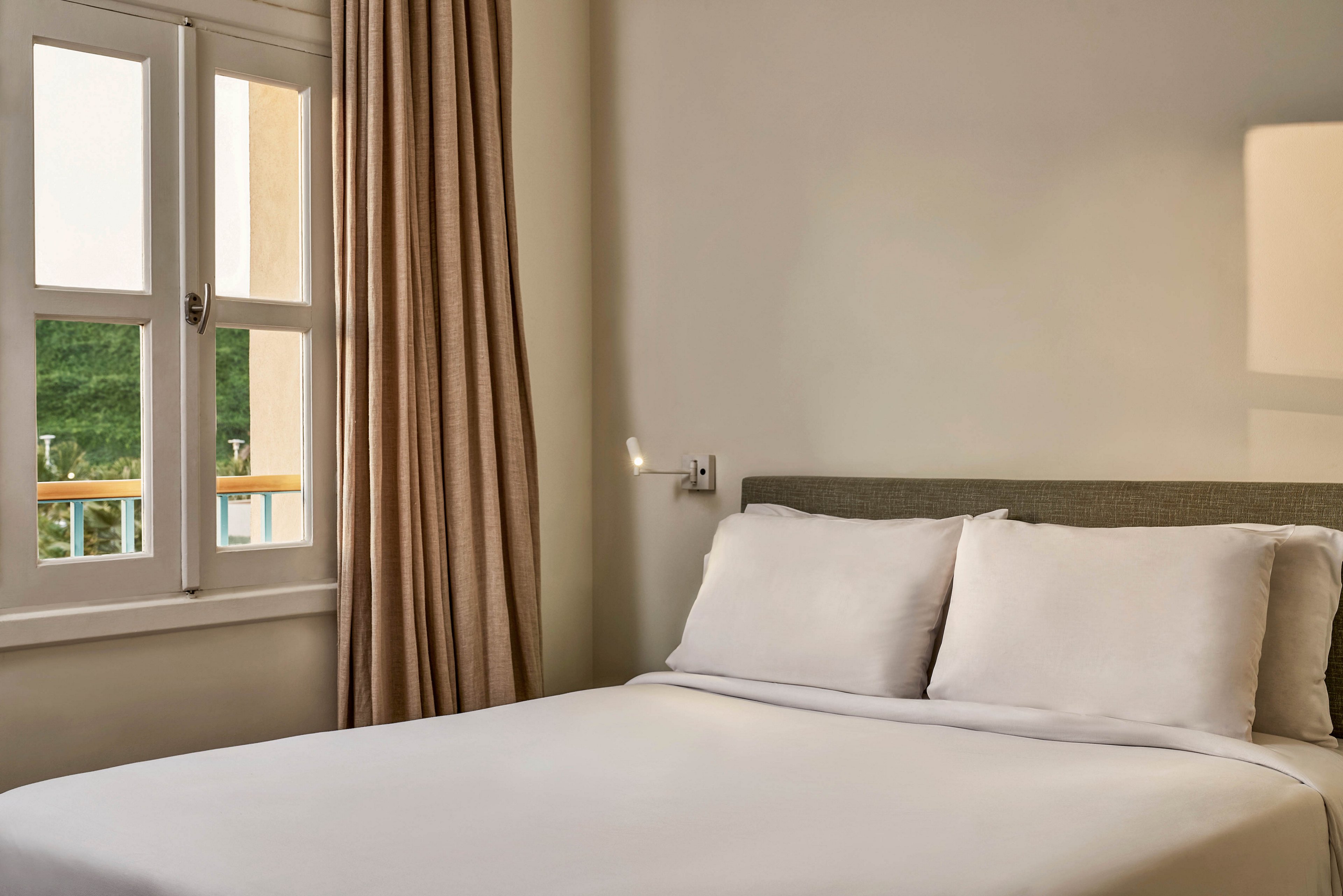 A neatly made bed with white linens is positioned next to a window, allowing natural sunlight to illuminate the room. Beige curtains hang beside the window, and a soft wall lamp adds a cozy touch to the minimalist decor. The scene conveys a calm and inviting atmosphere, with neutral tones and clean lines. No visible text or numbers are present in the image.