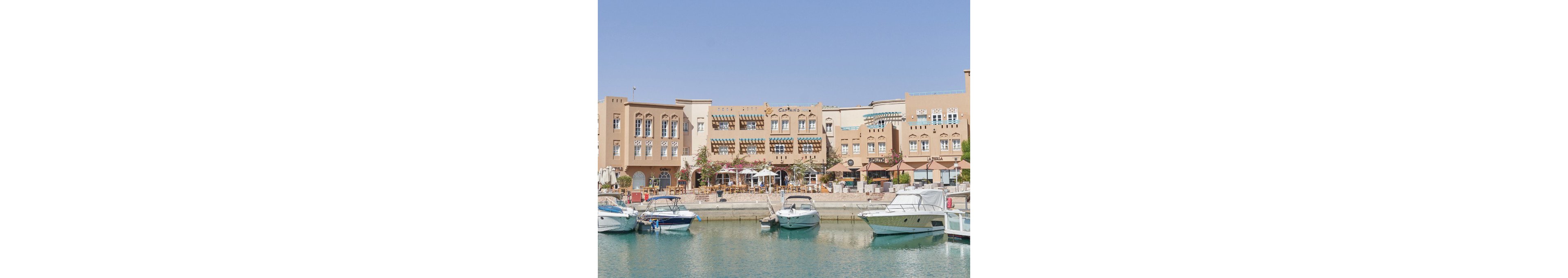 A row of small boats is docked in a calm marina in front of tan Mediterranean-style buildings. The scene features outdoor cafes and restaurants with shaded seating areas, and the clear blue sky enhances the relaxed, sunny atmosphere. The architecture includes arched windows, decorative awnings, and visible signage such as 'CLUB HOUSE.' The water reflects the boats and buildings, adding to the tranquil mood.