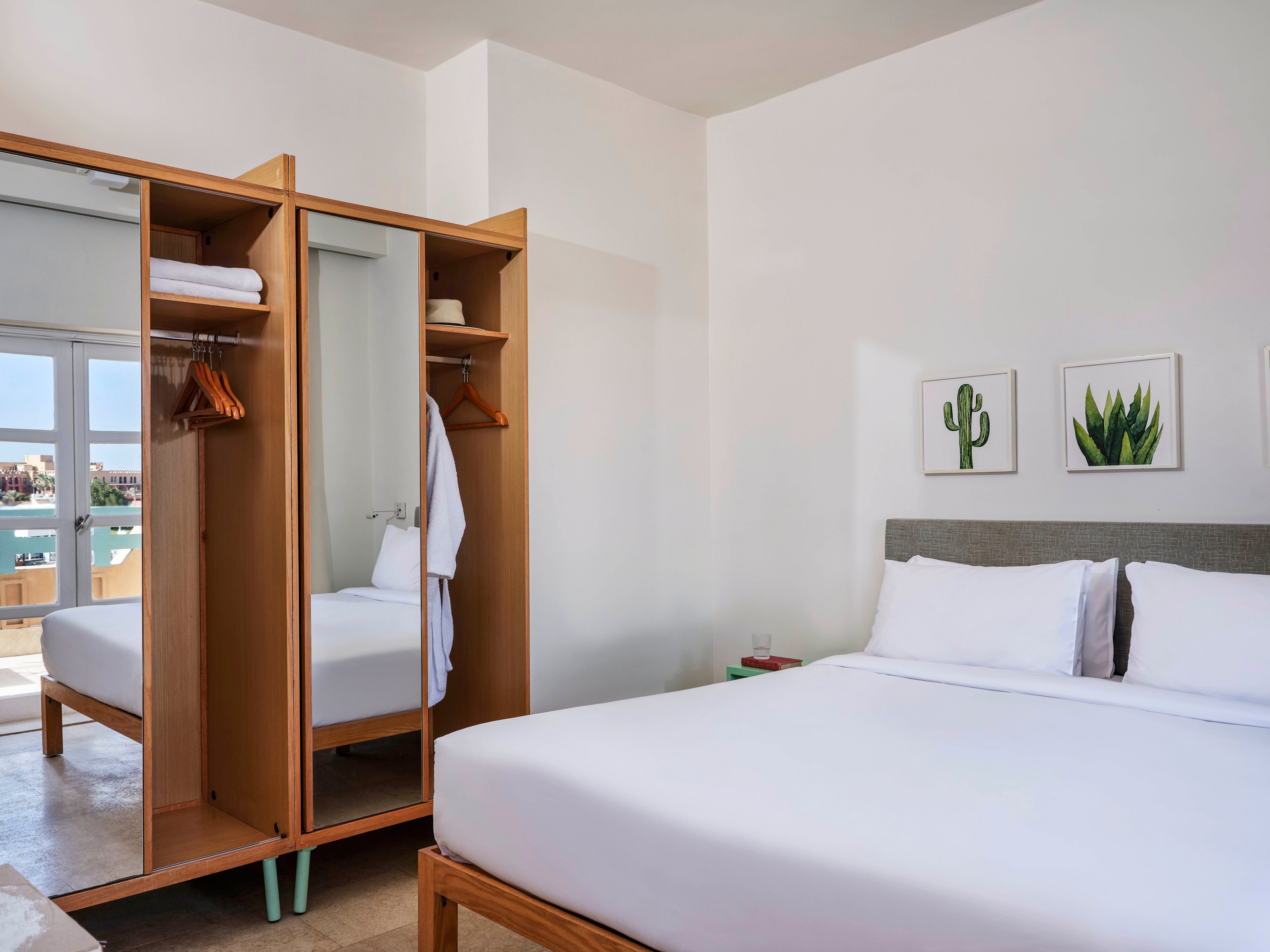 A contemporary hotel room features a neatly made bed with white linens and an open wooden wardrobe displaying hangers and a robe. The space is bright, with natural light streaming in from large windows. Two framed cactus prints decorate the wall above the bed, adding a touch of greenery to the minimalist decor. The overall palette is neutral with pops of green and wood tones.