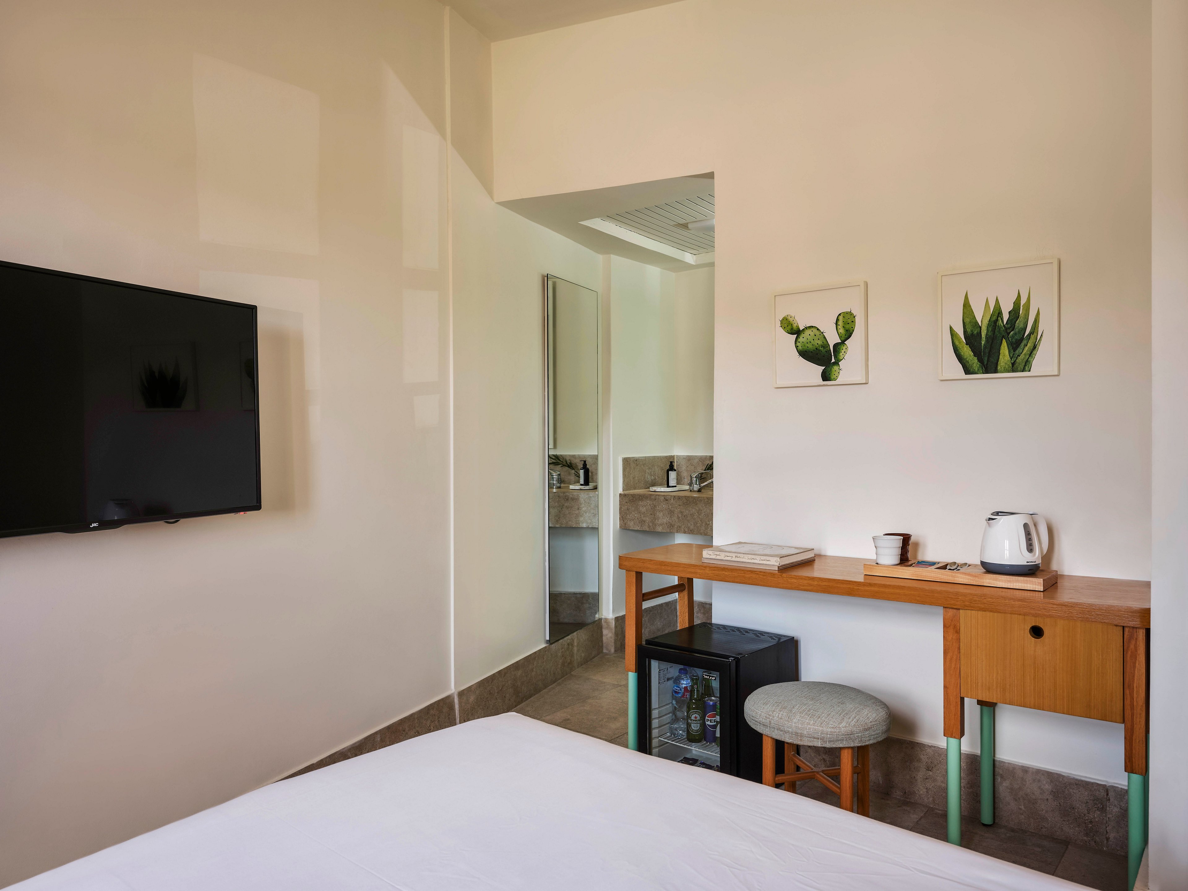 A contemporary hotel room features a wooden desk with a kettle, cup, and books, accompanied by a small stool. Two framed botanical prints hang above the desk, adding a touch of greenery to the minimalist decor. A flat-screen television is mounted on the wall, and a mini fridge is visible beneath the desk. The room is well-lit with neutral tones and clean lines, creating a calm and inviting atmosphere.