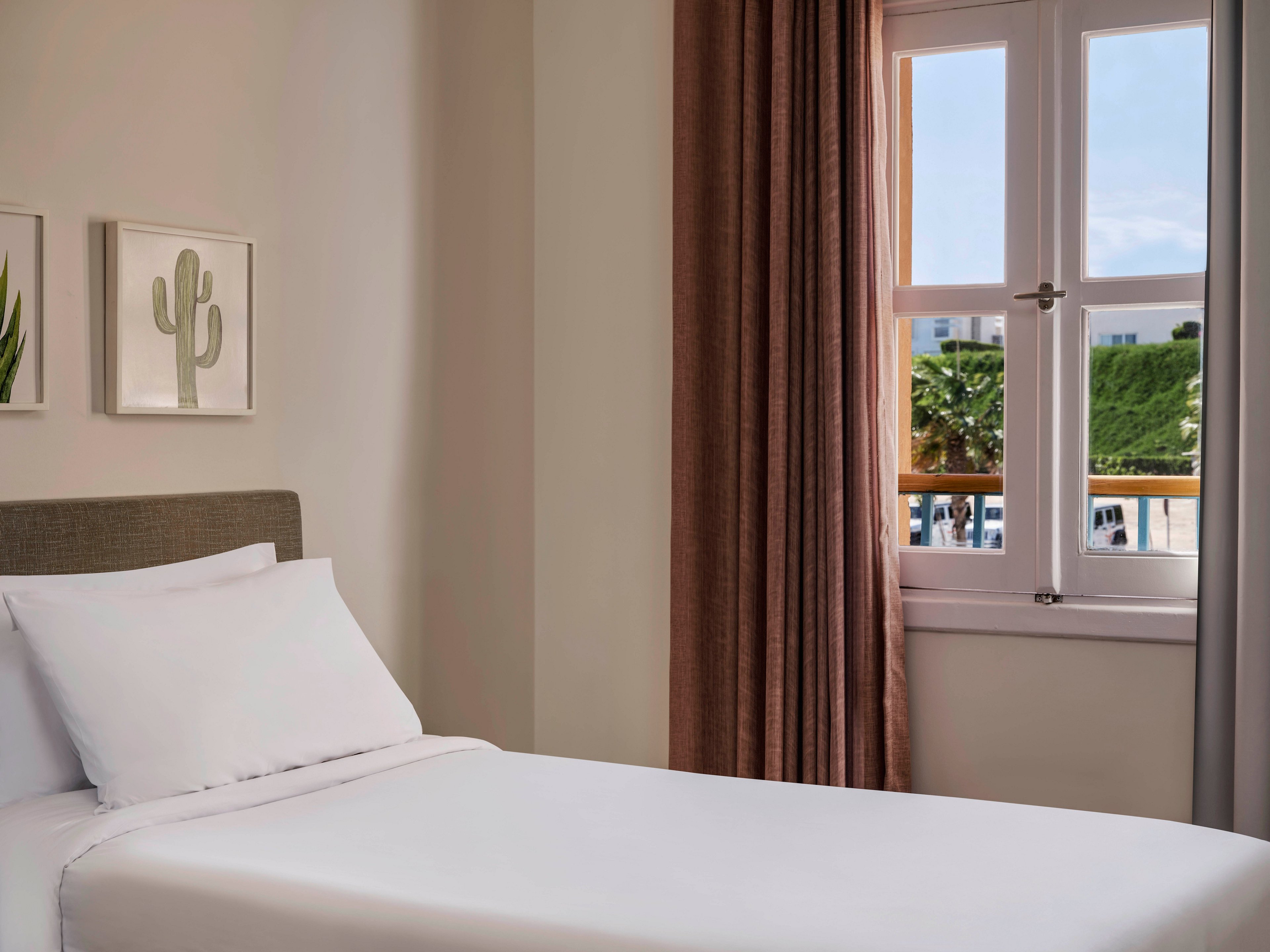 A neatly made bed with white linens is positioned beside a window with brown curtains. The room features minimalist decor, including framed cactus artwork on the wall. Outside the window, greenery and palm trees are visible, adding a touch of nature to the serene indoor setting. The palette is neutral and calming, emphasizing simplicity and comfort.