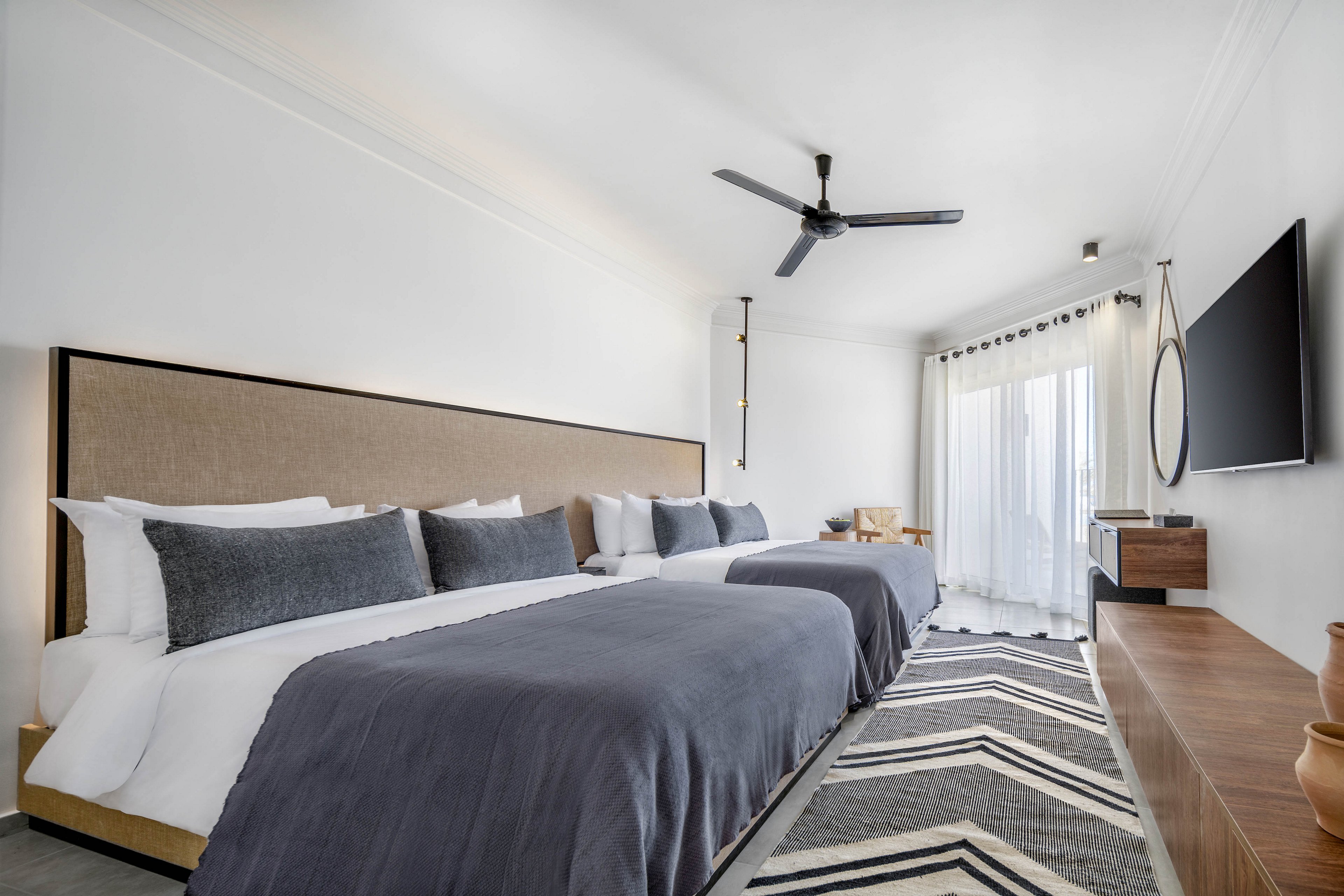 A spacious, contemporary hotel room features two large beds with gray bedding and multiple pillows. The room is decorated in a minimalist style with a neutral palette, accented by a geometric patterned rug and a wall-mounted flat-screen TV. Natural light streams in through sheer curtains covering a glass door that leads to a balcony. The setting is clean and inviting, with a ceiling fan and simple wooden furniture enhancing the modern aesthetic.