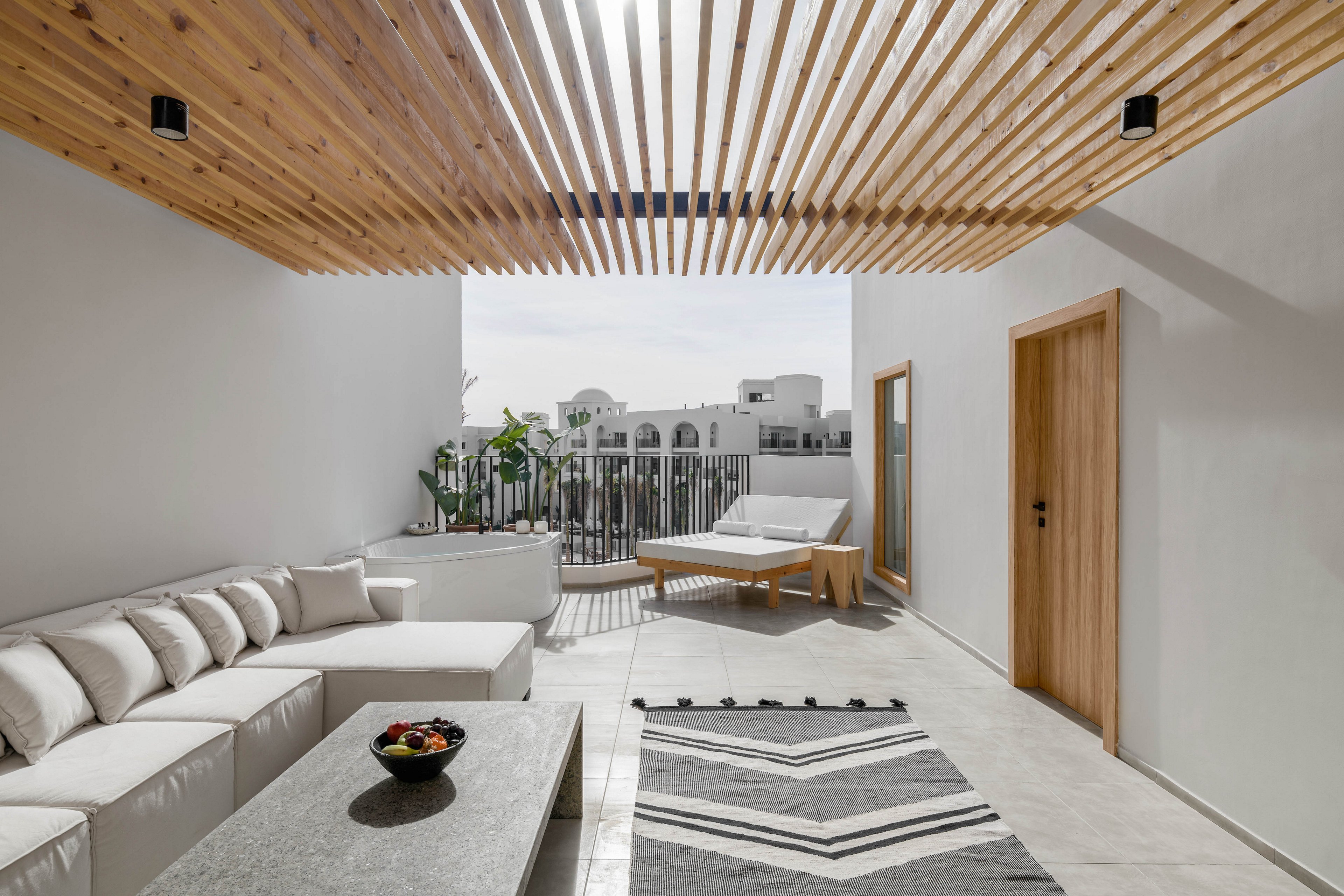 A contemporary outdoor lounge area features a white sectional sofa, a minimalist coffee table, and a geometric patterned rug. The space is shaded by a wooden slatted pergola, allowing natural light to filter through. In the background, a sunbed and potted plants add to the relaxing atmosphere, with white buildings visible beyond the balcony railing. The overall palette is neutral with natural wood accents, creating a calm and inviting mood.