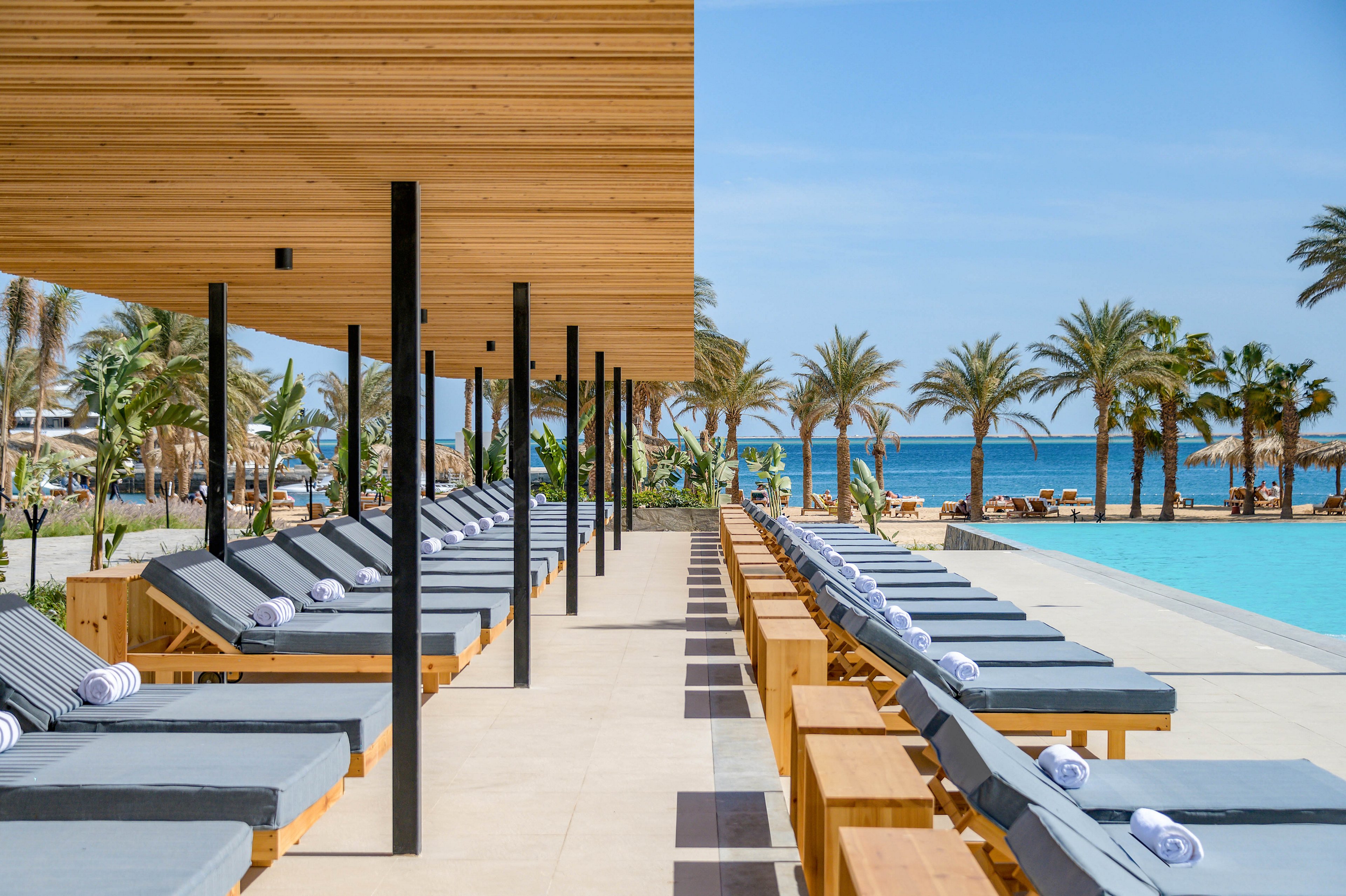 A row of contemporary lounge chairs with rolled towels lines a spacious poolside area under a wooden pergola. The setting overlooks a turquoise swimming pool and a scenic beach with palm trees, creating a tranquil and luxurious atmosphere. The sky is clear and blue, enhancing the relaxing vacation vibe. No visible text or numbers are present in the image.
