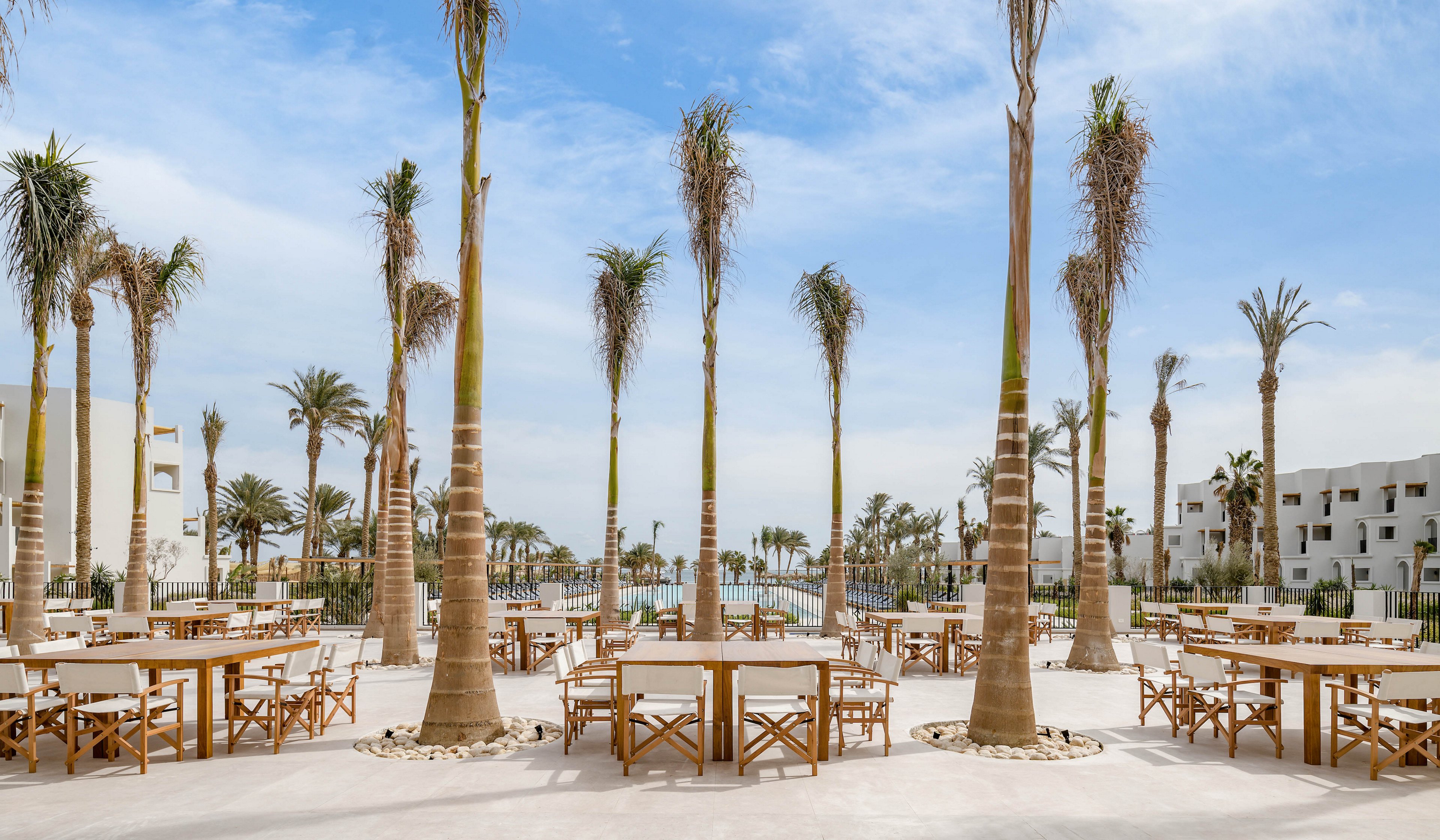 Spacious outdoor dining area features wooden tables and chairs arranged under tall palm trees. The setting is a modern resort with white buildings and a swimming pool visible in the background. The scene is bright and airy, with a clear blue sky and a relaxed, vacation-like atmosphere. No people are present, emphasizing tranquility and openness.