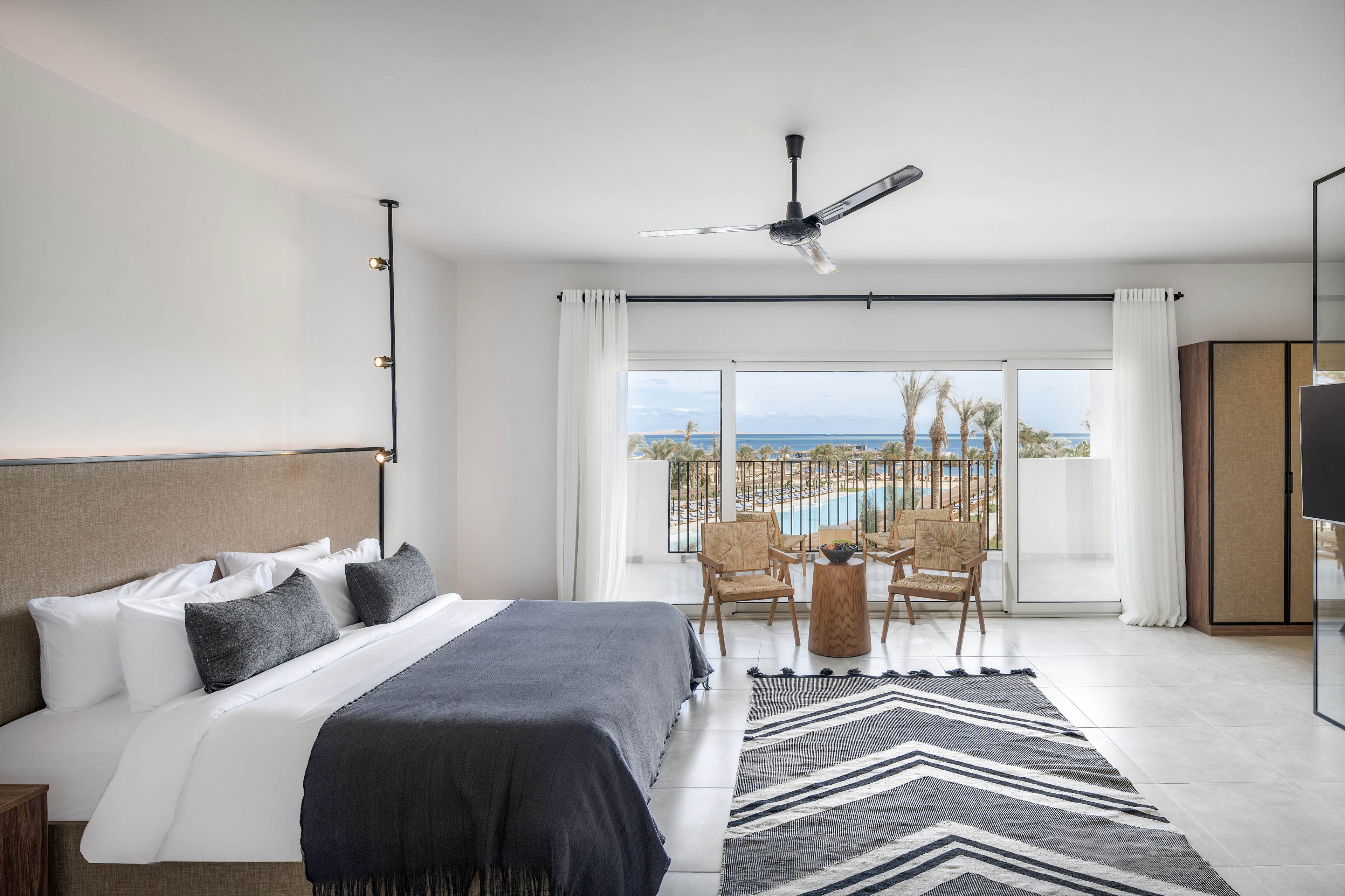 A spacious, contemporary hotel room features a large bed with white linens and dark accent pillows. The room opens to a balcony with two wicker chairs and a small table, overlooking a pool and the ocean beyond. Minimalist decor, a geometric rug, and neutral tones create a calm, inviting atmosphere. Natural light fills the space through wide glass doors, and a ceiling fan is centrally mounted.