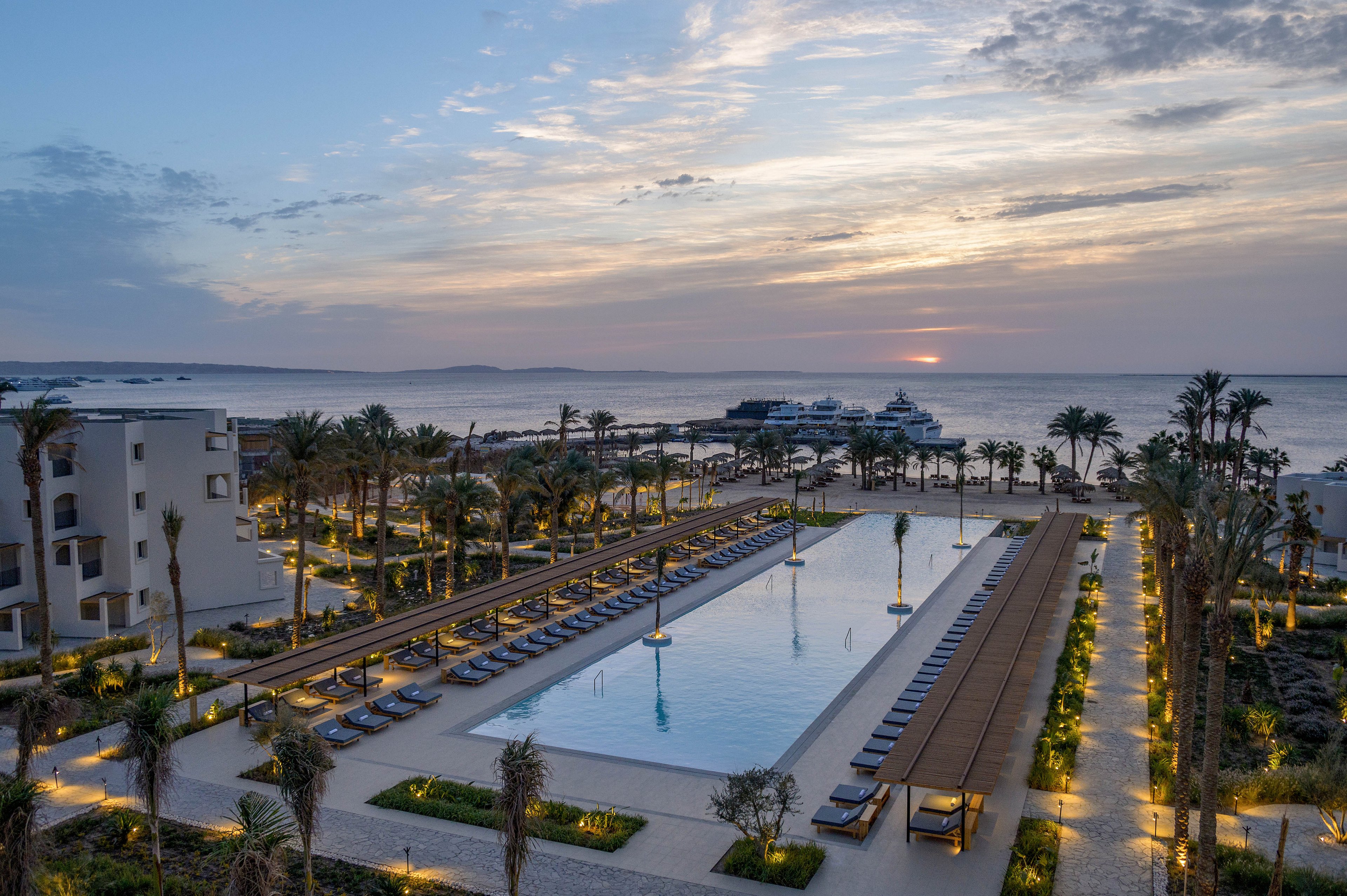 A large, rectangular swimming pool is the centerpiece of a luxury resort, surrounded by palm trees and lounge chairs. The setting is tranquil, with the sun setting over the ocean in the background, casting a warm glow across the scene. Modern buildings and landscaped pathways frame the pool, while yachts are visible on the water. The overall mood is serene and inviting, ideal for relaxation.