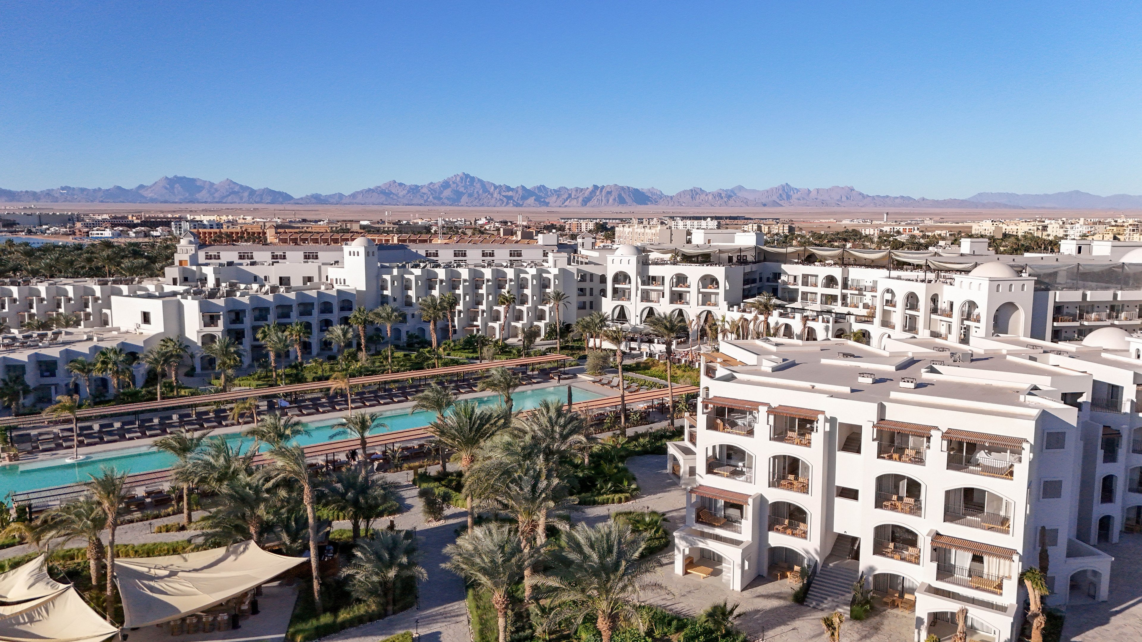 Aerial view of a large, modern resort complex featuring multiple white buildings and a long central swimming pool. The setting is lush with palm trees and landscaped gardens, with distant mountains visible under a clear blue sky. The architecture is Mediterranean-inspired, and the atmosphere is serene and inviting. No visible text or numbers are present in the image.