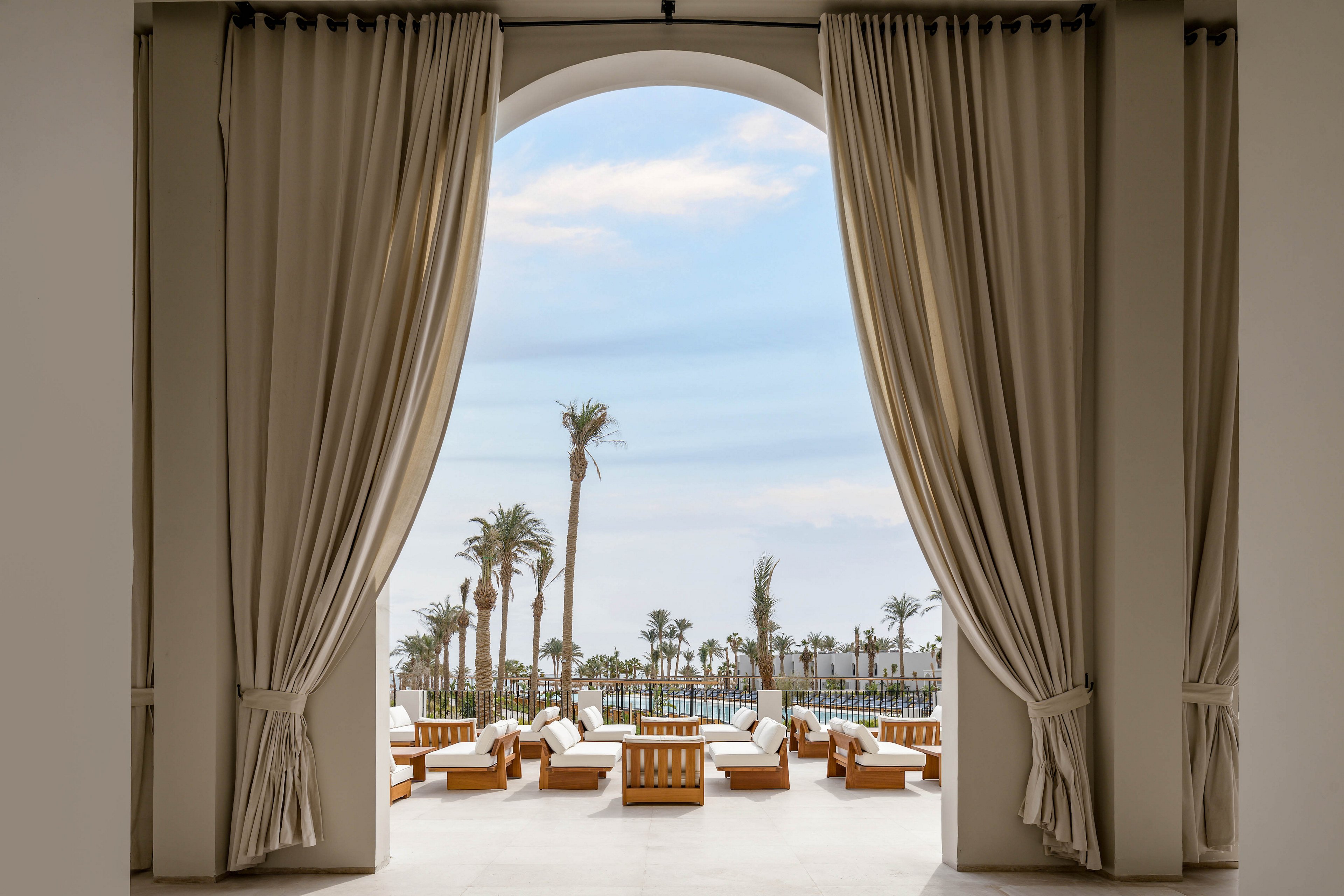A spacious lounge area opens to a scenic outdoor pool framed by tall palm trees. Elegant beige curtains and wooden lounge chairs create a relaxed, upscale atmosphere. The setting is tranquil, with soft natural light and a clear blue sky visible through the large archway. The scene suggests a tropical or coastal resort environment, ideal for relaxation and leisure.