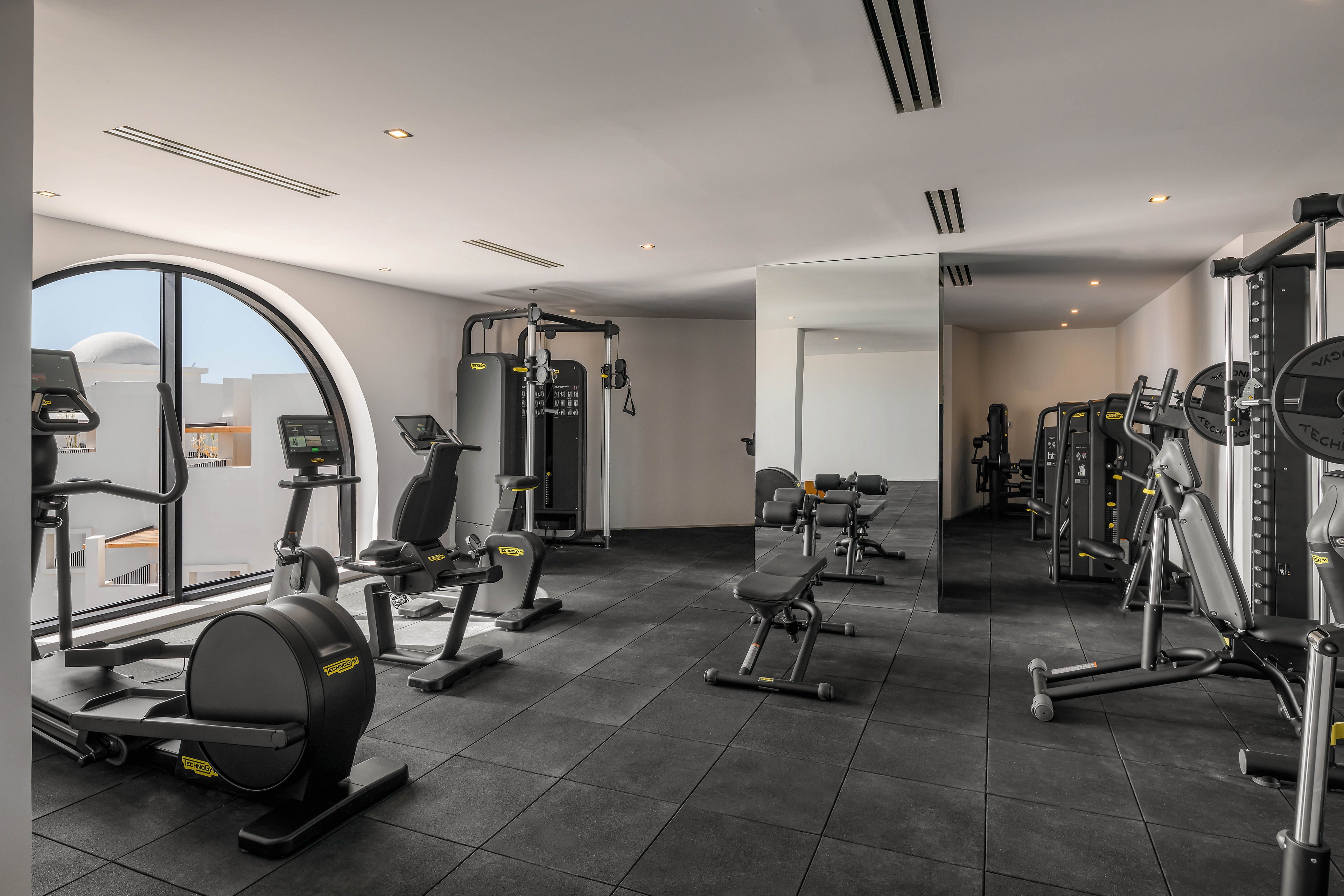 A spacious gym features a variety of exercise machines, including treadmills, stationary bikes, and weight benches. The room is well-lit with natural light from a large arched window and has a clean, minimalist design. Black flooring and mirrored walls enhance the contemporary atmosphere, while the equipment is neatly arranged for easy access.