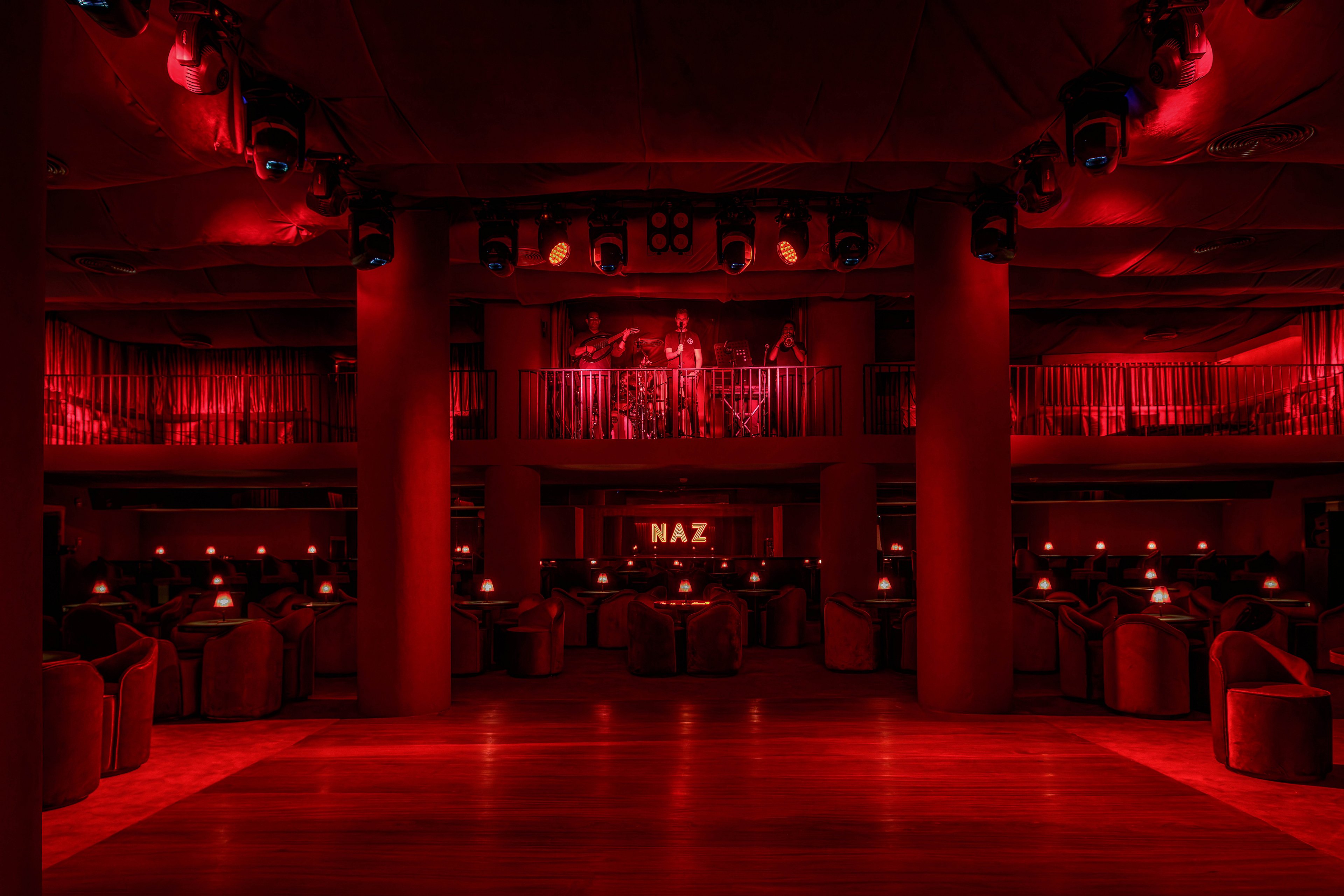 A nightclub interior is illuminated with dramatic red lighting, creating a moody and atmospheric setting. A live band performs on a balcony stage above the main floor, which is filled with round tables and plush seating. The word 'NAZ' is clearly visible in illuminated letters at the back of the venue. The overall ambiance is intimate and vibrant, emphasizing nightlife and entertainment.