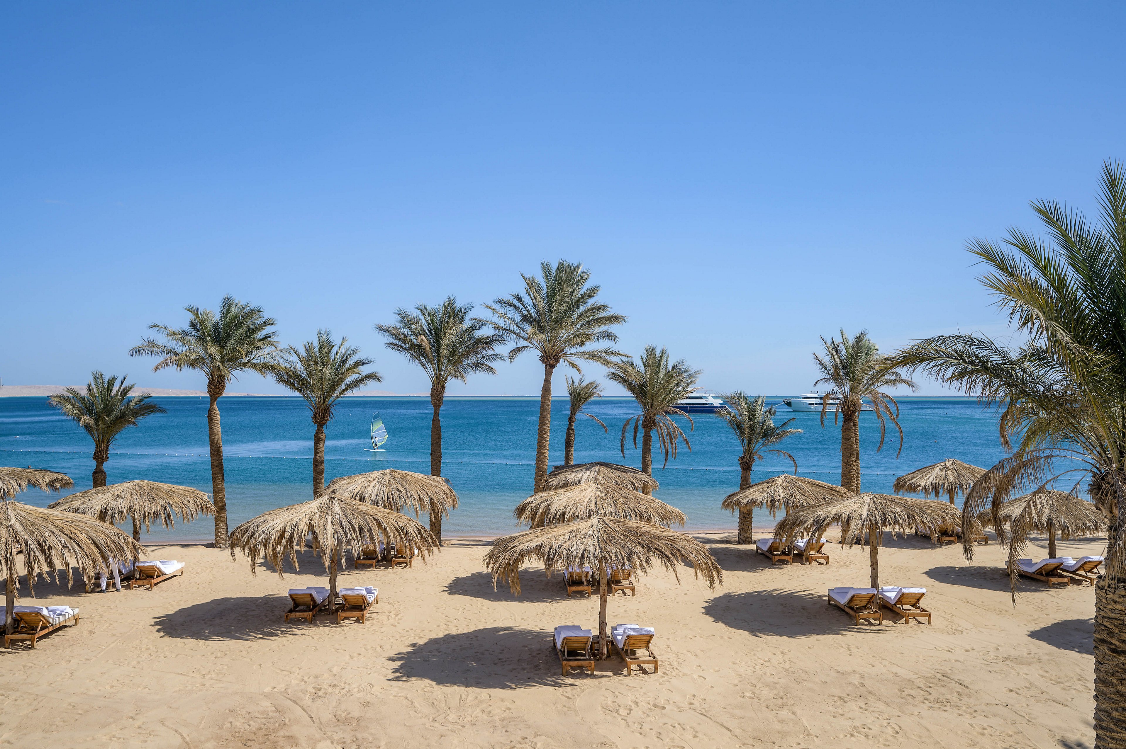 A serene sandy beach is lined with palm trees and straw umbrellas, each shading a pair of lounge chairs. The clear blue sea stretches into the horizon under a bright sky, with a windsurfer and boats visible in the distance. The scene evokes a relaxing, vacation atmosphere with natural tones and a tranquil mood.