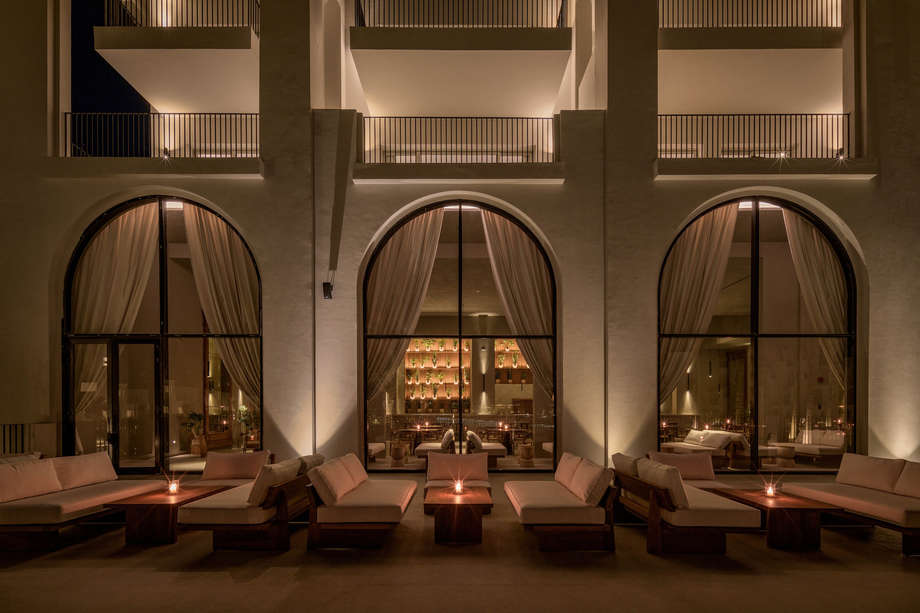 A sophisticated outdoor lounge area is set against the backdrop of a modern hotel facade at night. Large arched windows reveal a warmly lit interior with a stylish bar and plush seating. The scene features soft ambient lighting, neutral tones, and an inviting atmosphere, perfect for evening relaxation or social gatherings.
