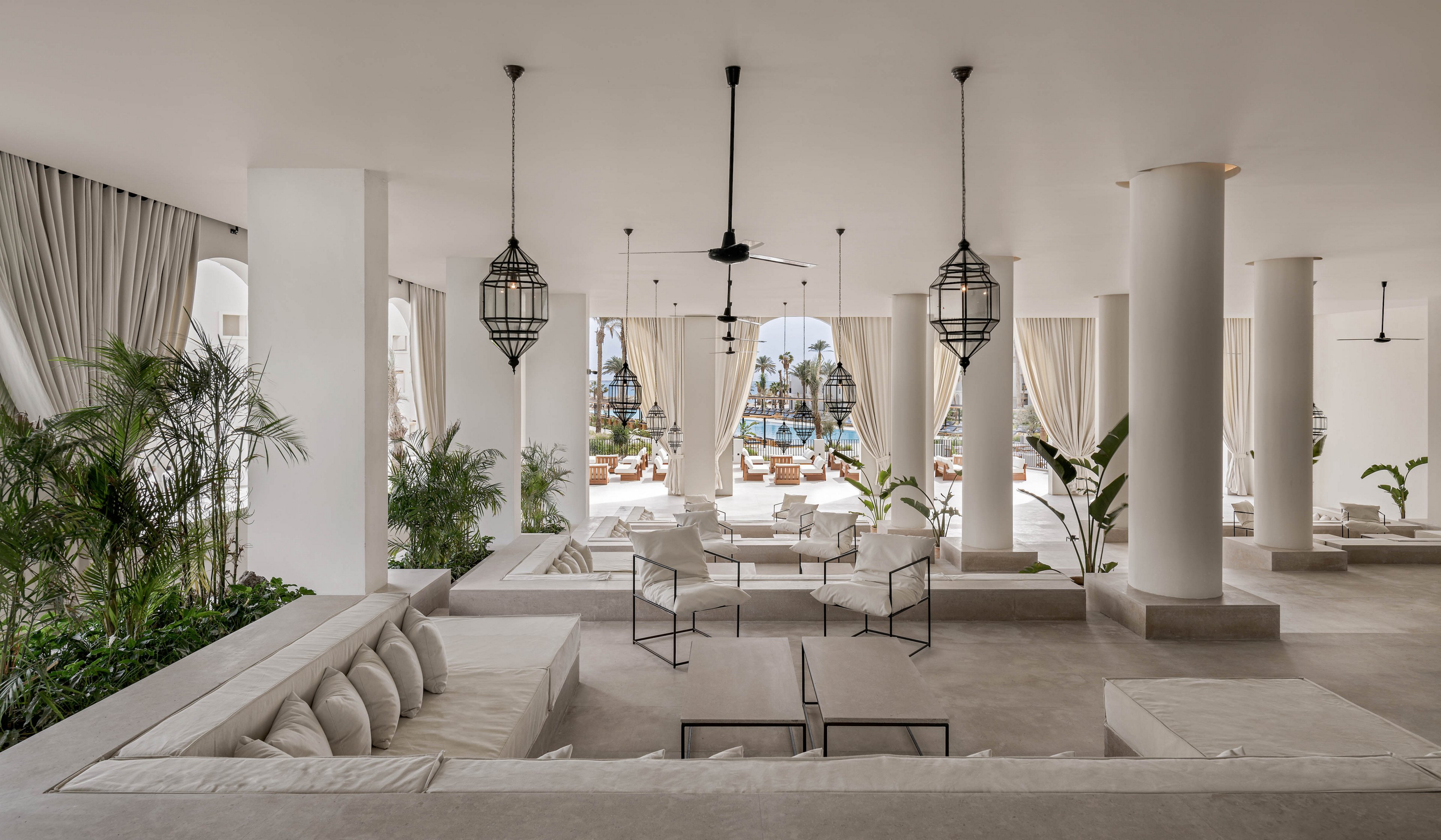 A spacious, contemporary lounge area features white cushioned seating, minimalist chairs, and large columns. The setting is open-air with flowing curtains, tropical plants, and hanging lanterns, creating a serene and inviting atmosphere. Natural light floods the space, and a pool with palm trees is visible in the background. The overall palette is neutral, emphasizing relaxation and luxury.