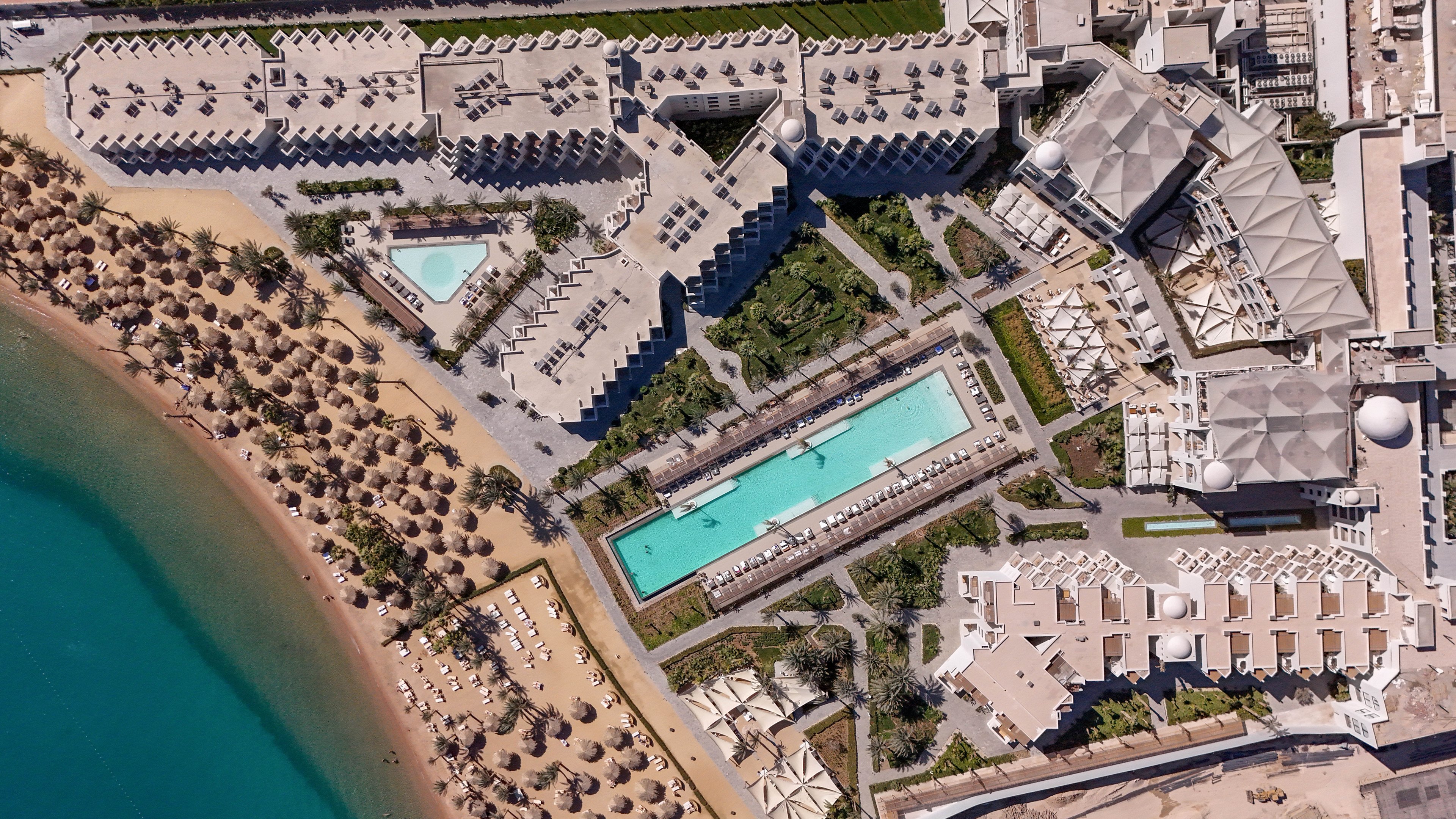 This aerial photograph captures a sprawling luxury resort complex situated along a sandy beachfront. The image highlights multiple swimming pools, including a large rectangular pool and a triangular pool, surrounded by sun loungers and palm-thatched umbrellas. The turquoise shoreline, geometric architecture, and lush landscaping create a vibrant and inviting atmosphere. No visible text or numeric values are present in the image.