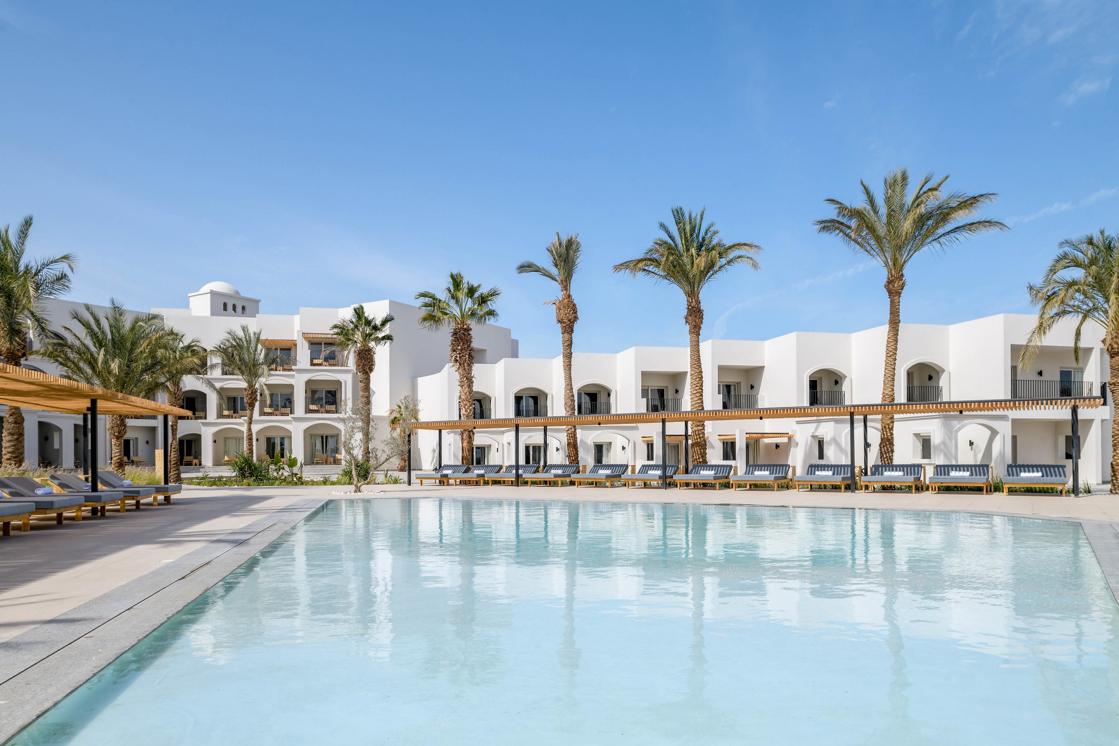 A contemporary white hotel building surrounds a large outdoor swimming pool under a clear blue sky. Tall palm trees line the poolside, and shaded lounge chairs are arranged neatly along the deck. The setting is calm and inviting, evoking a relaxed vacation atmosphere with minimalist architecture and natural elements.