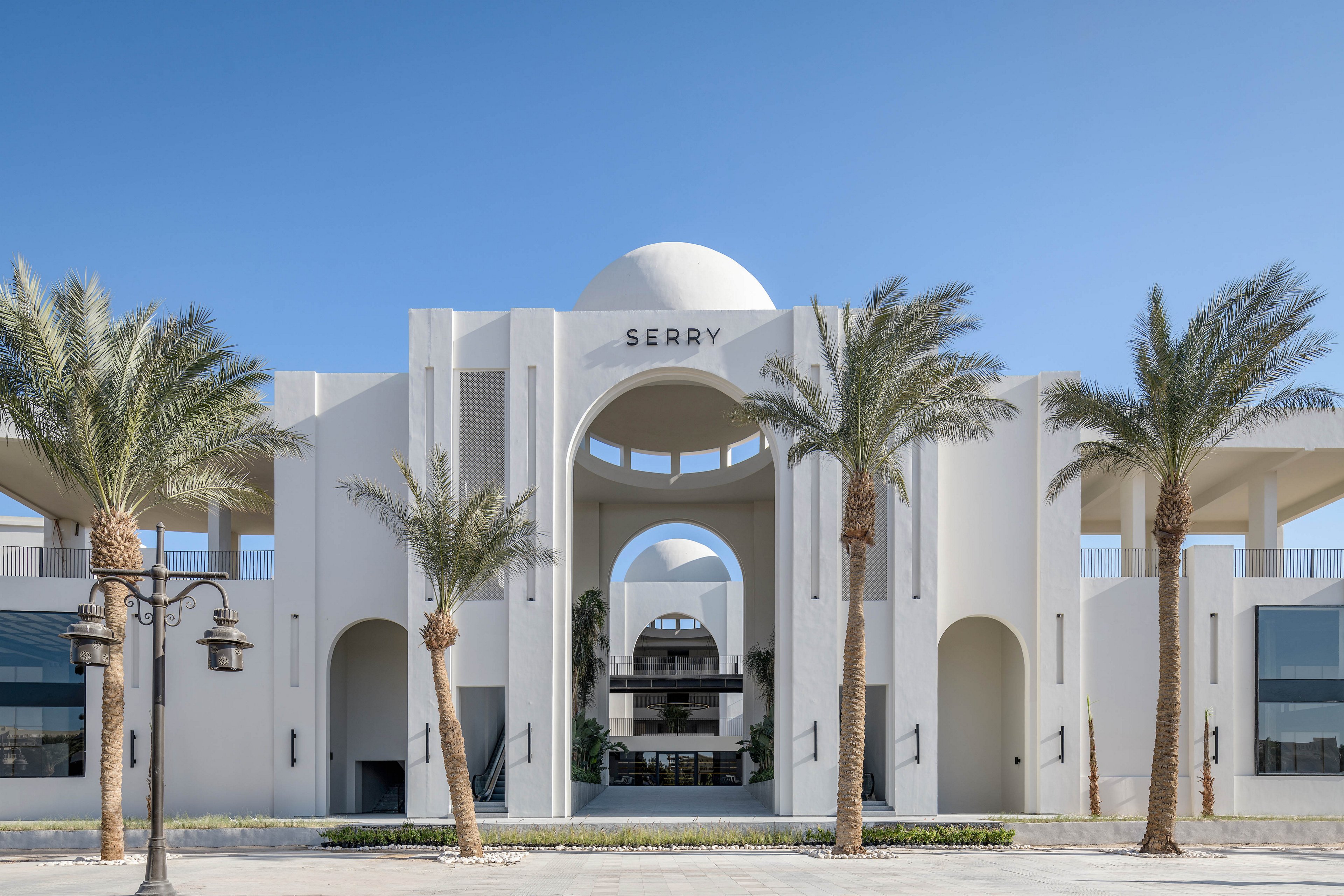A contemporary hotel building features a striking white facade with large geometric arches and a central dome. The word 'SERRY' is clearly visible above the main entrance. Tall palm trees line the front, and a vintage-style street lamp stands to the left. The scene is set under a clear blue sky, emphasizing the clean lines and Mediterranean-inspired architecture.