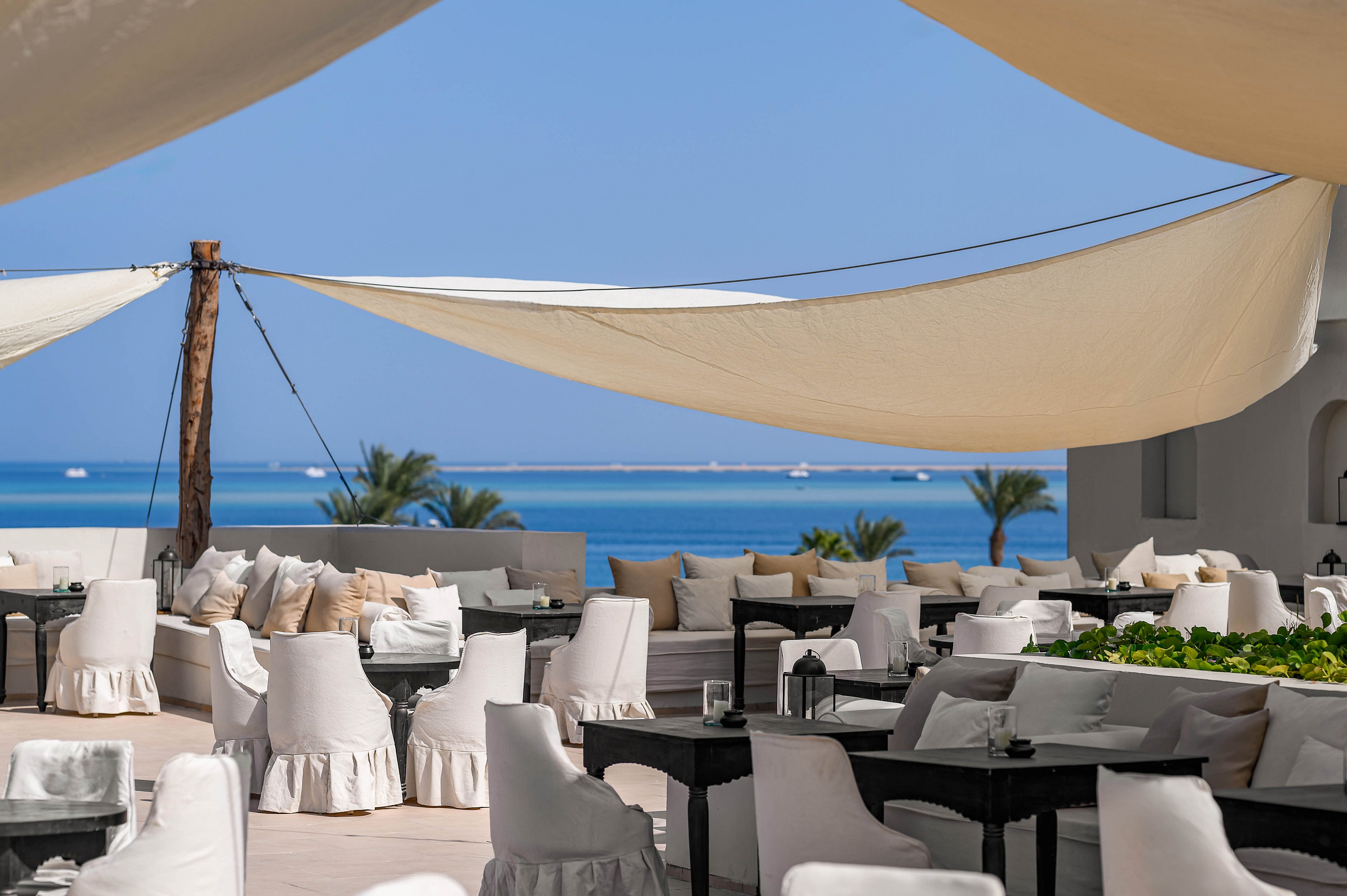 A stylish outdoor seating area features white fabric-covered chairs and sofas arranged under large canvas canopies. The setting overlooks a serene blue ocean with palm trees and clear skies in the background. The atmosphere is calm and inviting, with a neutral color palette and modern decor. No visible text or numbers are present in the image.