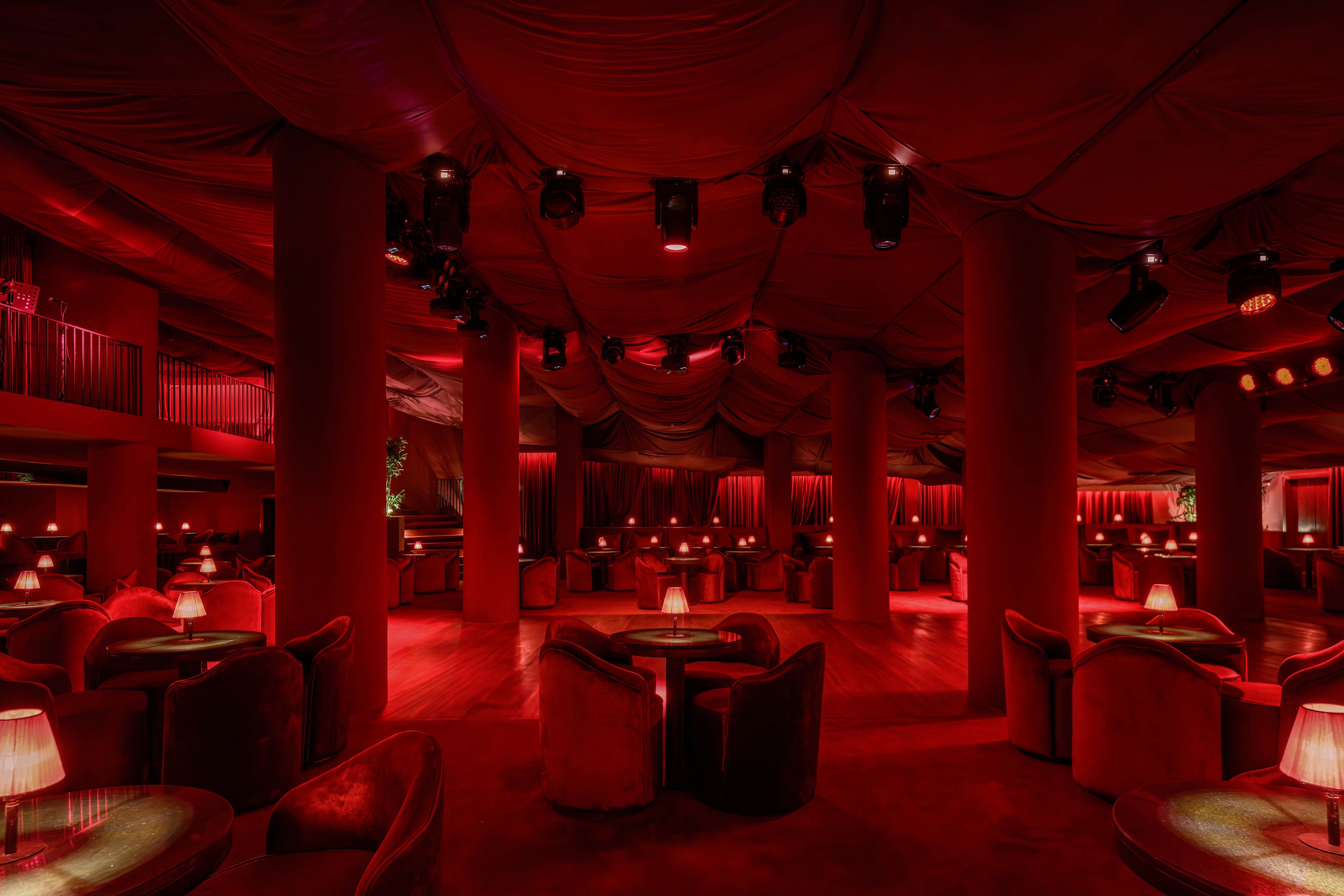 A spacious, upscale lounge features plush seating arranged around small tables, all illuminated by soft red lighting. The setting is intimate and sophisticated, with draped ceilings and numerous spotlights overhead. The room is empty, emphasizing the moody, atmospheric vibe and the elegant arrangement of furniture. No visible text, logos, or numeric values are present in the image.