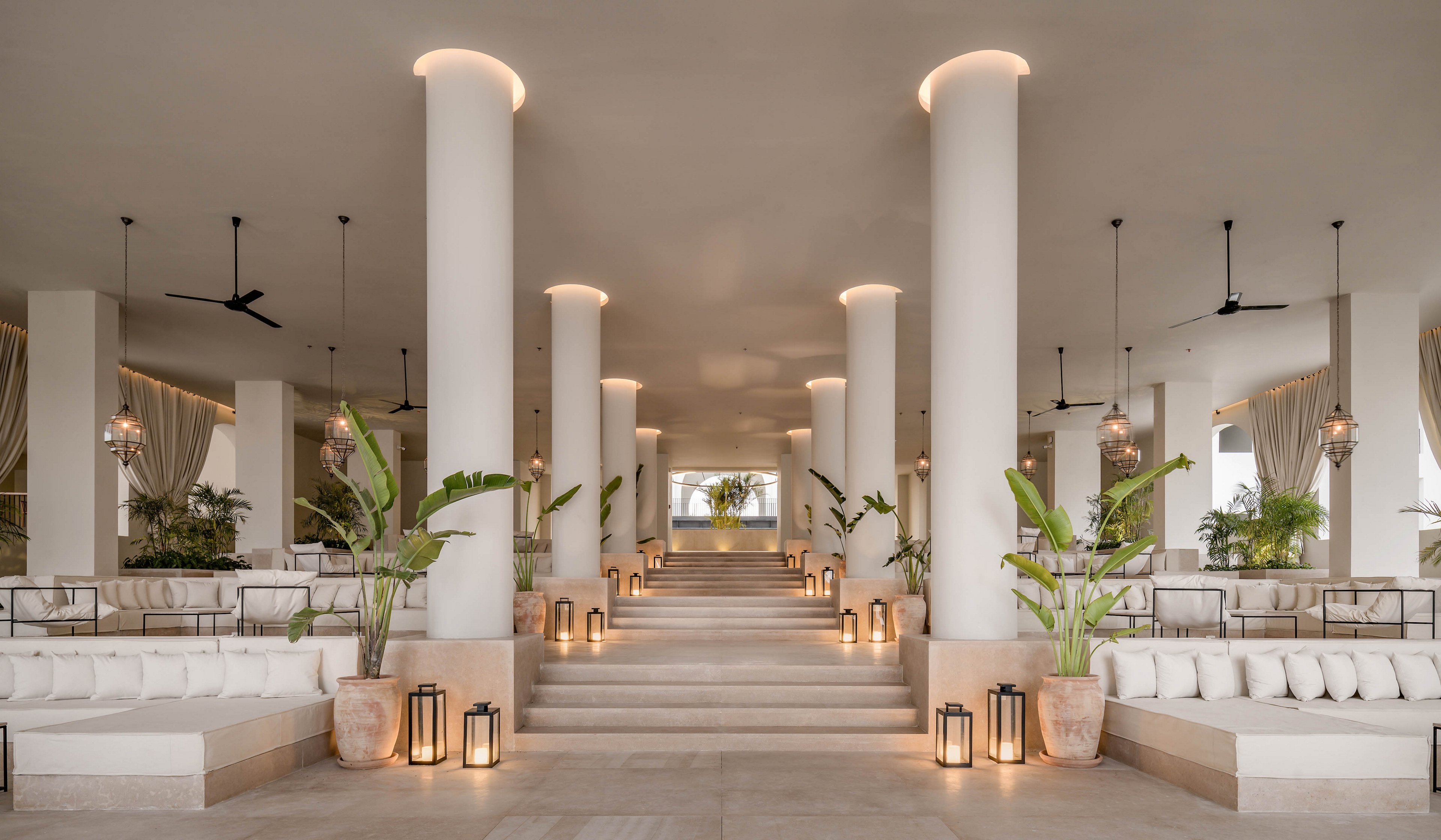 A spacious, contemporary hotel lobby features large white columns and neutral-toned seating areas. The setting is open-air with natural light, potted tropical plants, and decorative lanterns lining the steps. The mood is serene and inviting, with soft ambient lighting and minimalist decor. Ceiling fans and draped curtains add to the relaxed, resort-like atmosphere.