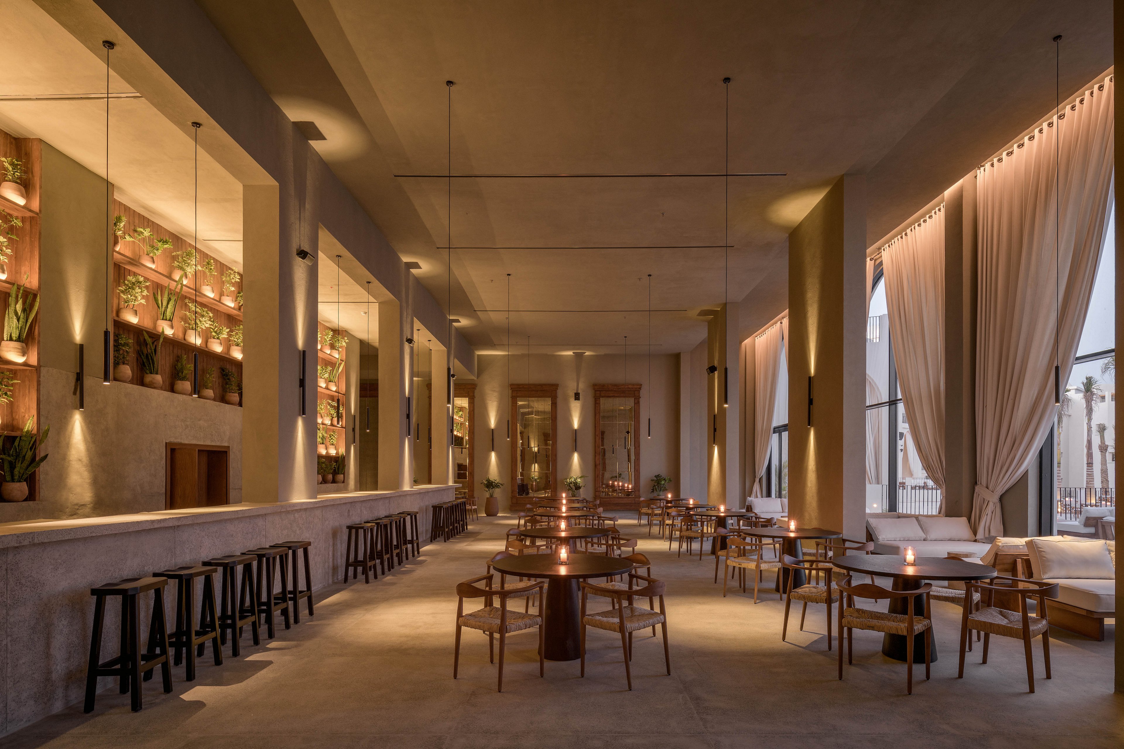 A spacious, contemporary restaurant features minimalist wooden tables and chairs, with a long bar lined with stools on one side. Soft, warm lighting and neutral tones create an inviting and elegant atmosphere. Vertical shelves with potted plants add a touch of greenery, while large windows with sheer curtains let in natural light. The setting is empty, emphasizing the tranquil and sophisticated mood.