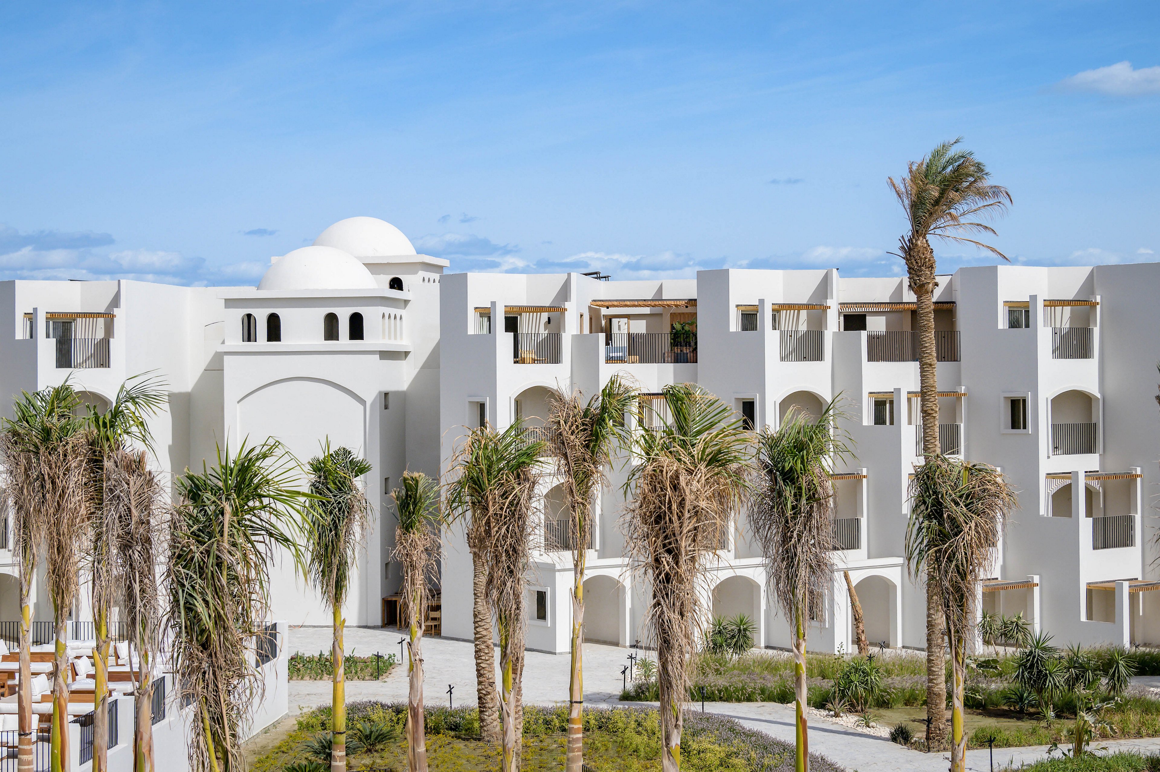 A contemporary white hotel building features arched windows and domed rooftops, set against a clear blue sky. The foreground is lined with tall palm trees and a manicured garden, evoking a Mediterranean or North African resort atmosphere. The architecture is characterized by clean lines, geometric shapes, and a bright, sunlit palette.