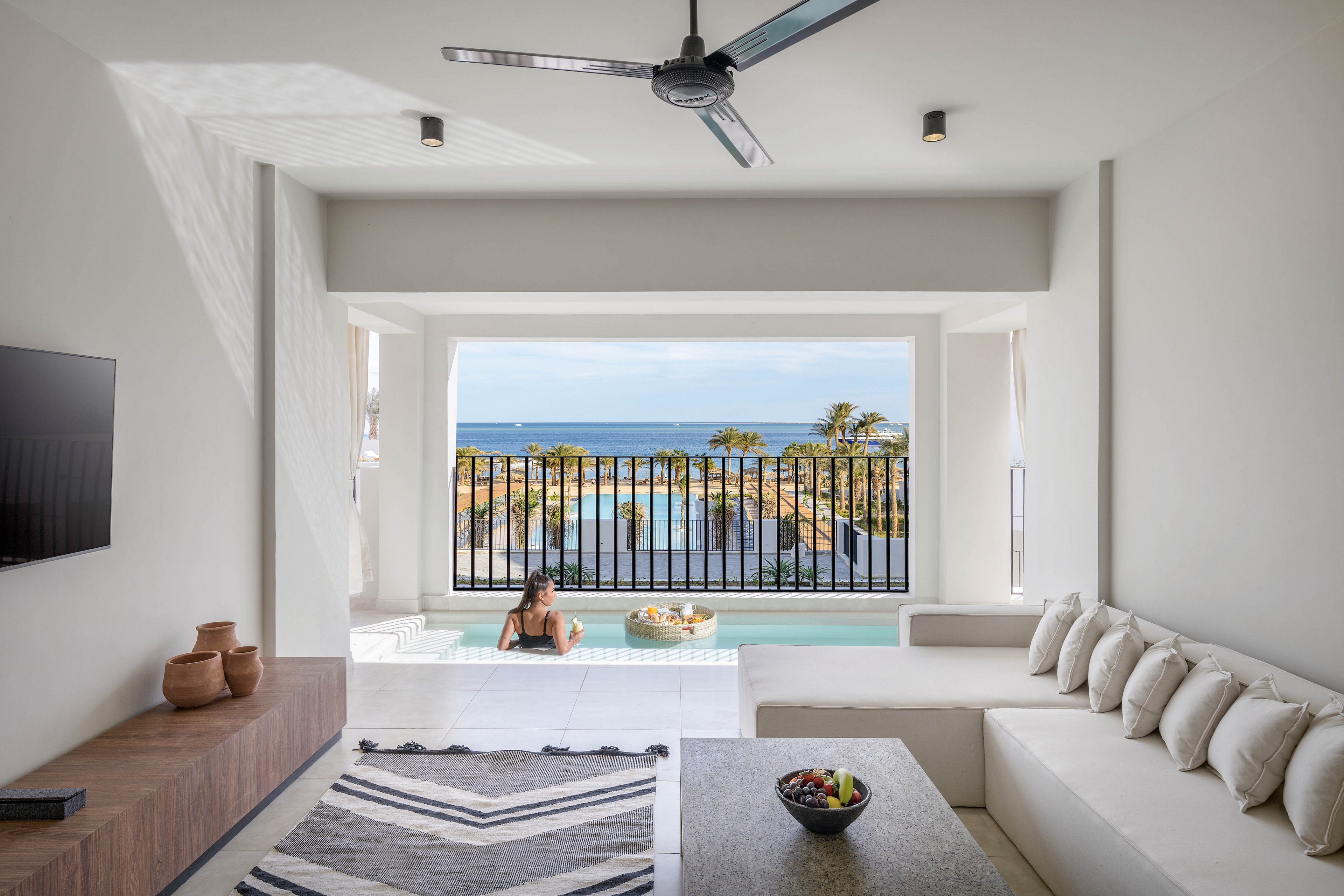 A contemporary living space features a spacious white sectional sofa, geometric rug, and minimalist decor. Through the open terrace, a woman relaxes in a private plunge pool overlooking palm trees and the ocean. The setting is bright and airy, with neutral tones and natural light enhancing the tranquil, luxurious atmosphere.