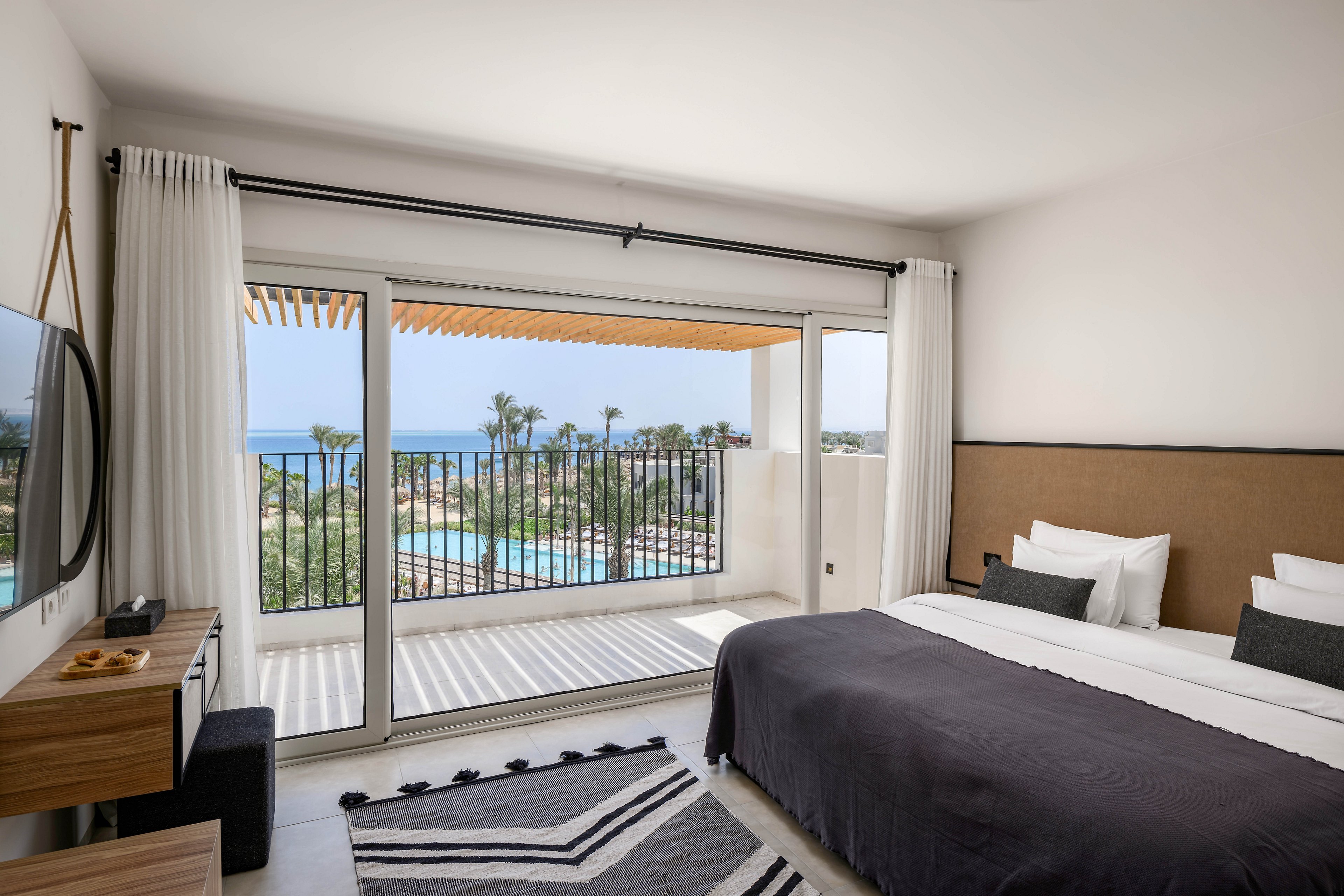 A contemporary hotel room features a large bed with crisp white linens and a dark throw blanket. The space opens to a private balcony overlooking a pool, palm trees, and the ocean beyond. Minimalist decor, natural light, and a monochrome rug create a calm, inviting atmosphere. The room includes a wall-mounted TV and a wooden desk, with no visible text or numbers present.