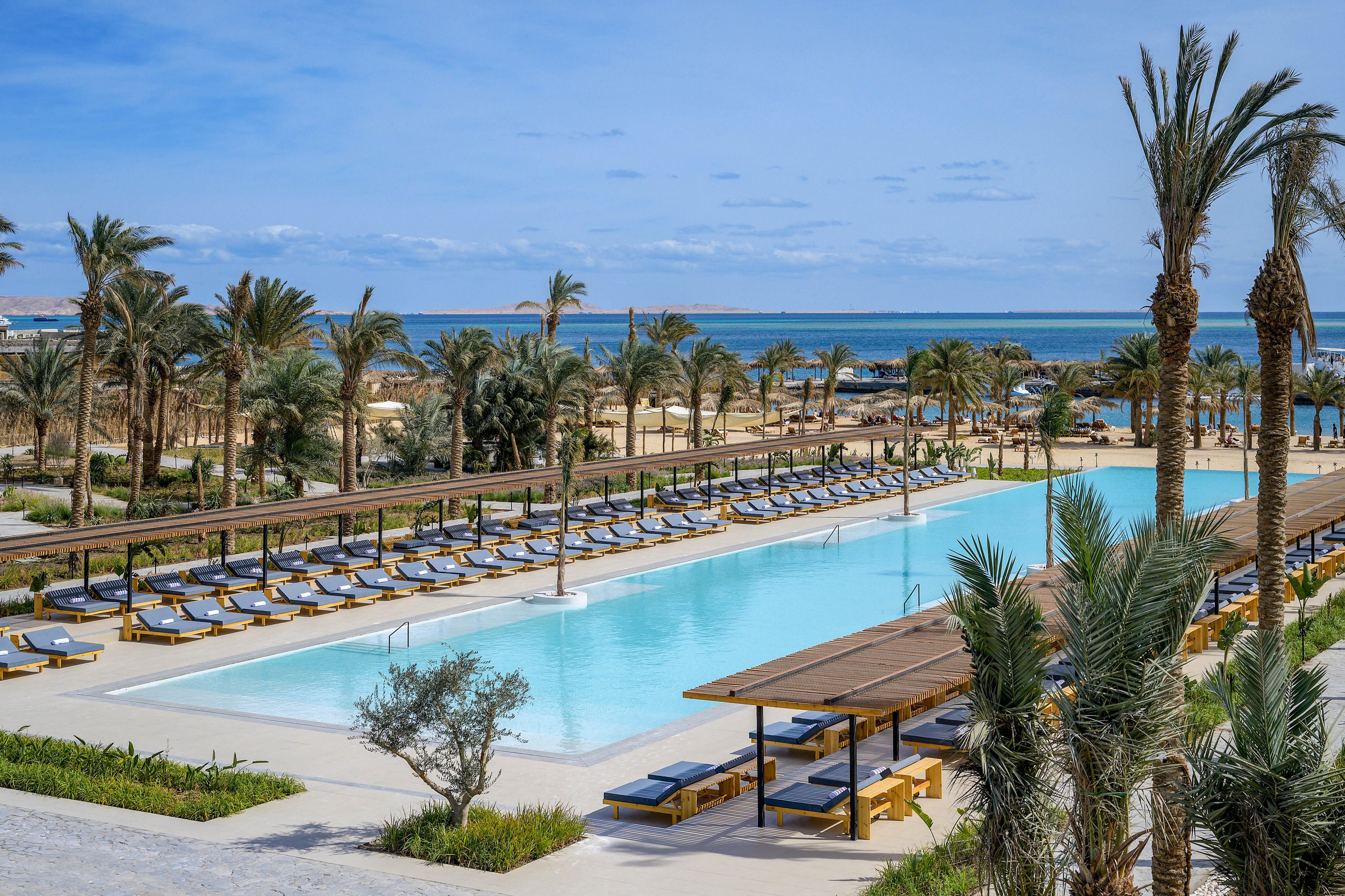 A large, rectangular swimming pool is surrounded by rows of sun loungers and shaded cabanas, set within a tropical resort landscape. Tall palm trees frame the pool area, with the ocean visible in the background under a partly cloudy sky. The tranquil setting suggests a relaxing vacation atmosphere, with clean lines and a modern design aesthetic. No visible people or text are present in the image.