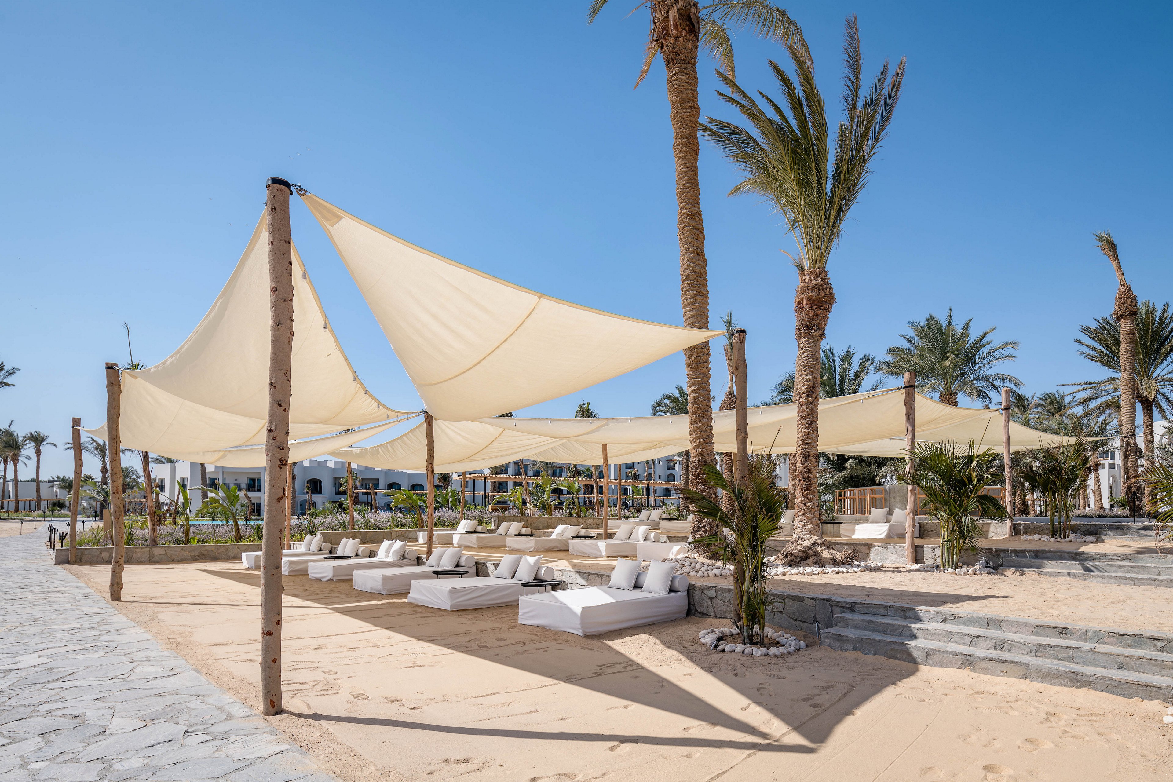 A stylish outdoor lounge area is set up on a sandy beach, featuring white cushioned seating under large fabric sunshades. Tall palm trees surround the space, creating a relaxed and inviting atmosphere. The sky is clear and blue, and the overall palette is neutral with natural tones. No visible text or numbers are present in the image.