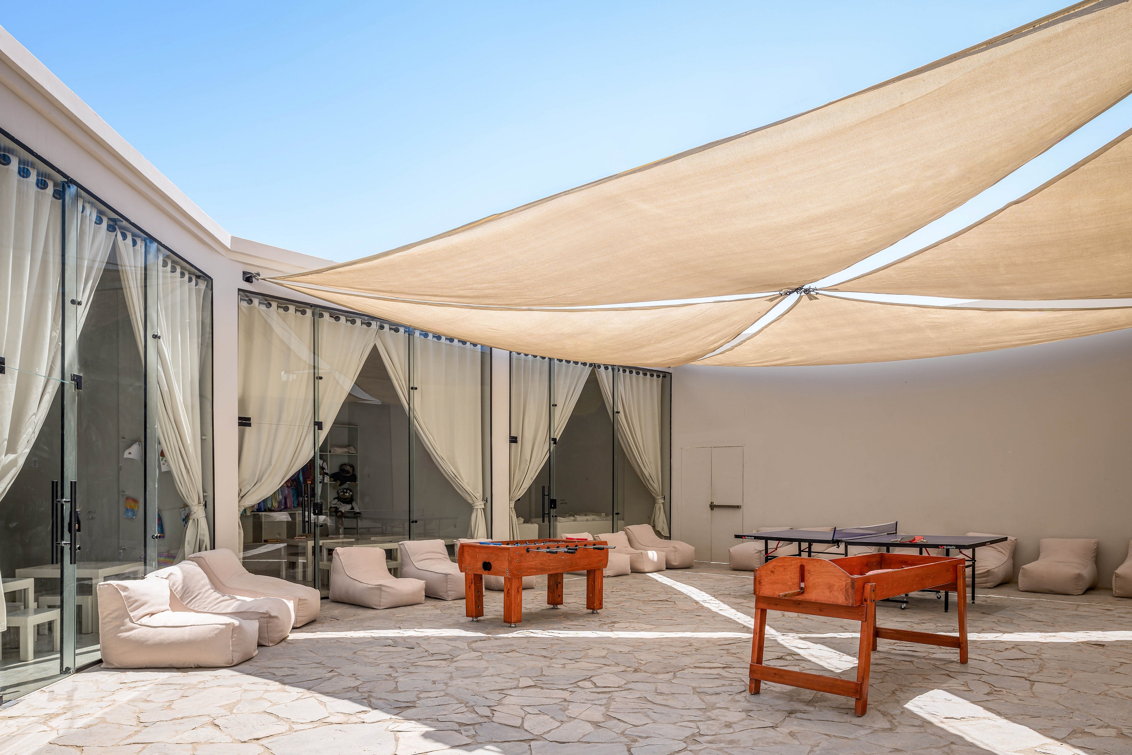 A contemporary outdoor lounge area features comfortable bean bag chairs and glass walls with draped curtains. Two wooden foosball tables and a table tennis setup are arranged on a stone patio under large beige shade sails. The setting is bright and airy, ideal for relaxation and social activities.