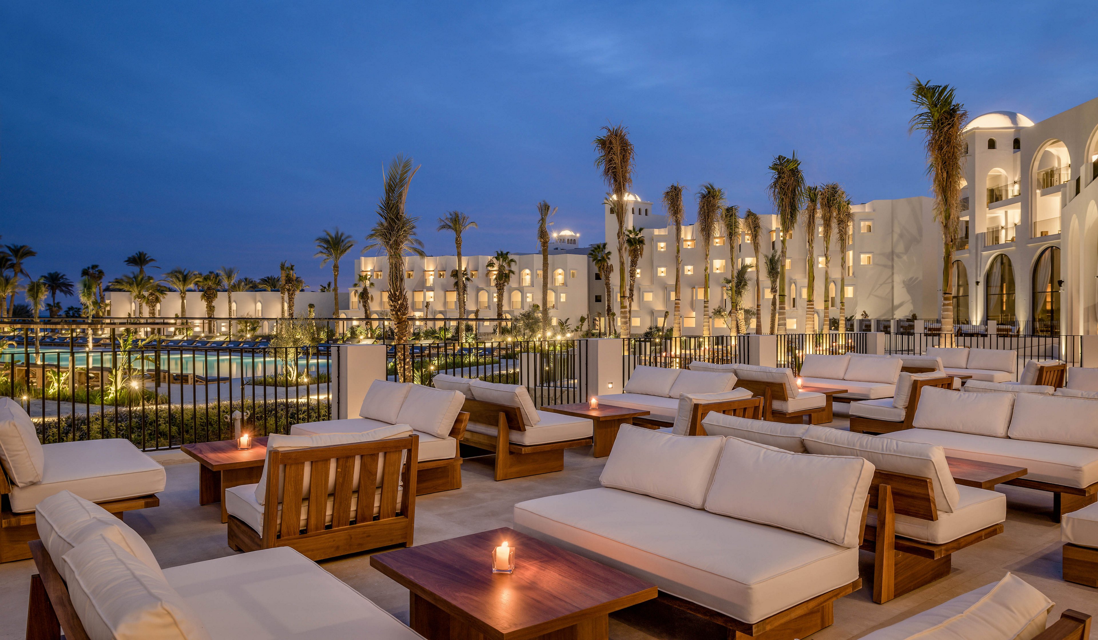 Spacious outdoor seating area with plush white sofas and wooden tables is set against a backdrop of a modern resort. The scene is illuminated by soft candlelight and ambient evening lighting, creating a tranquil and inviting atmosphere. Palm trees and a large pool are visible, enhancing the luxurious vacation vibe. The architecture features white facades and arched windows, contributing to a Mediterranean or Middle Eastern style.