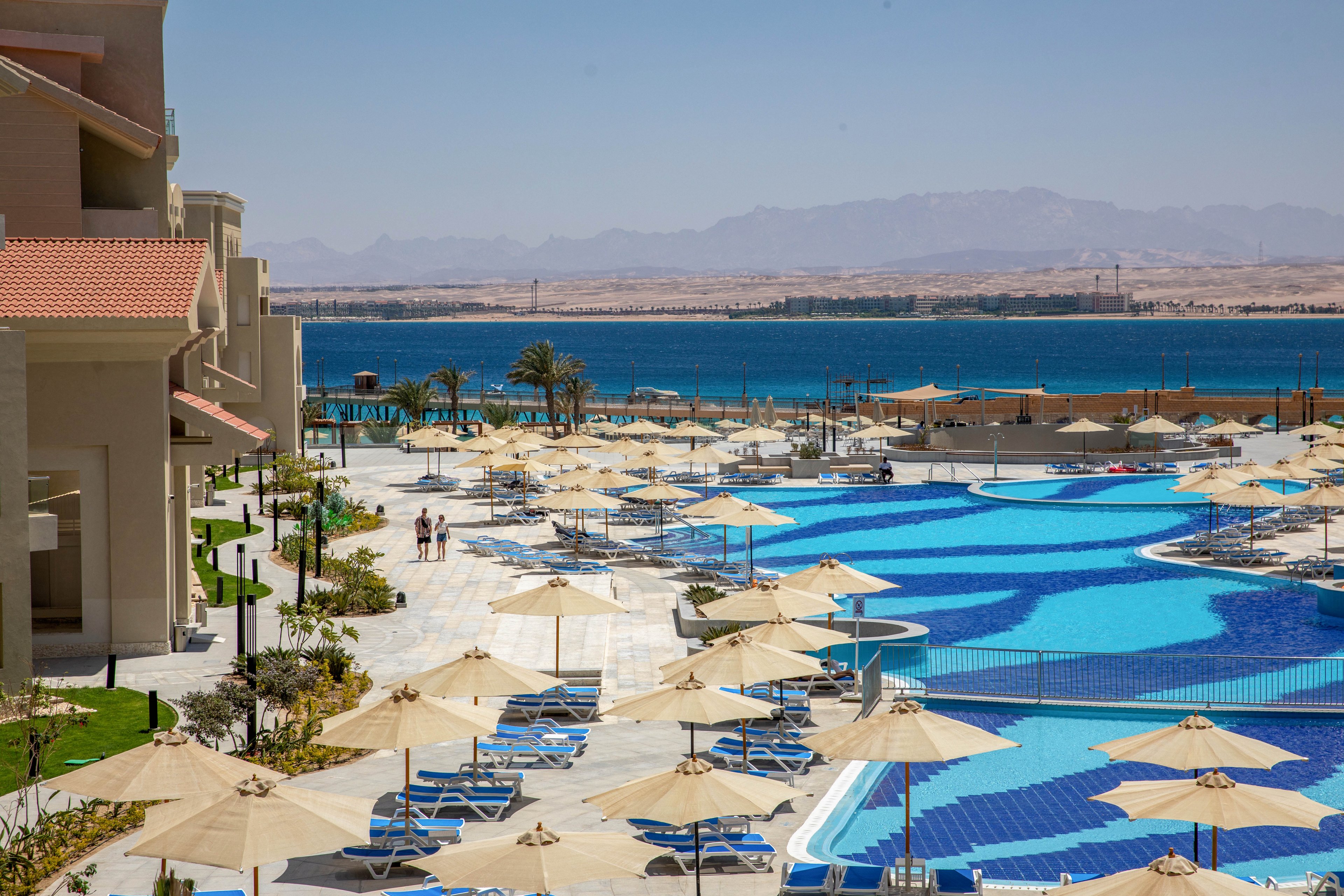 A modern resort features a large, winding swimming pool surrounded by sun loungers and umbrellas. The setting overlooks a vibrant blue sea with distant mountains under a clear sky. A few people are seen walking along the poolside, enhancing the relaxed vacation atmosphere. The architecture is contemporary with beige facades and red-tiled roofs. The overall mood is tranquil and inviting, ideal for leisure and relaxation.