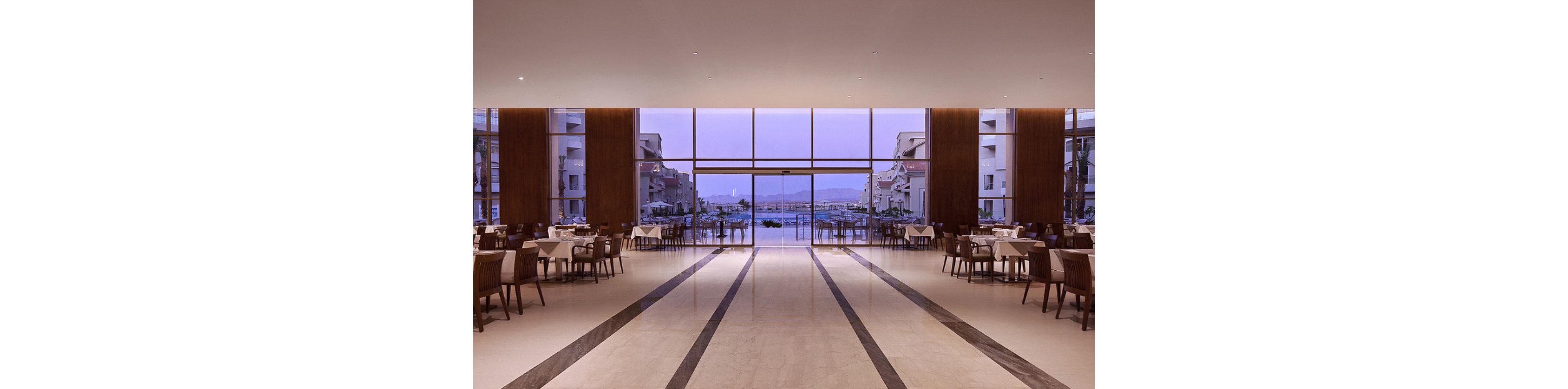 A large, elegant restaurant interior features neatly arranged tables and chairs on a polished marble floor. Floor-to-ceiling windows reveal an outdoor pool area and resort buildings, with a soft purple evening light illuminating the space. The setting is calm and inviting, with a minimalist, contemporary design and a tranquil atmosphere.