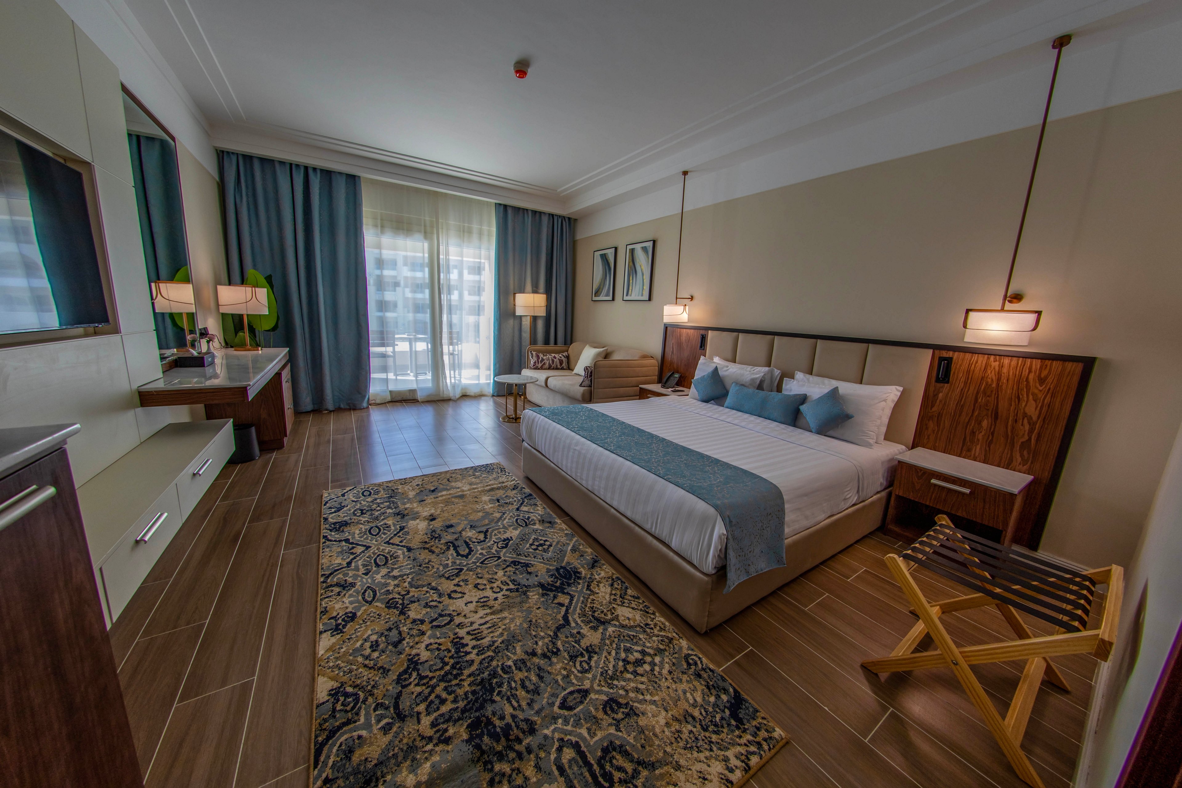 A spacious, contemporary hotel room features a king-sized bed with blue and white bedding, flanked by wooden nightstands and pendant lights. The room includes a patterned area rug, a work desk, and a sitting area near large windows with sheer curtains. The decor is modern with neutral tones and blue highlights, creating a calm and inviting atmosphere.