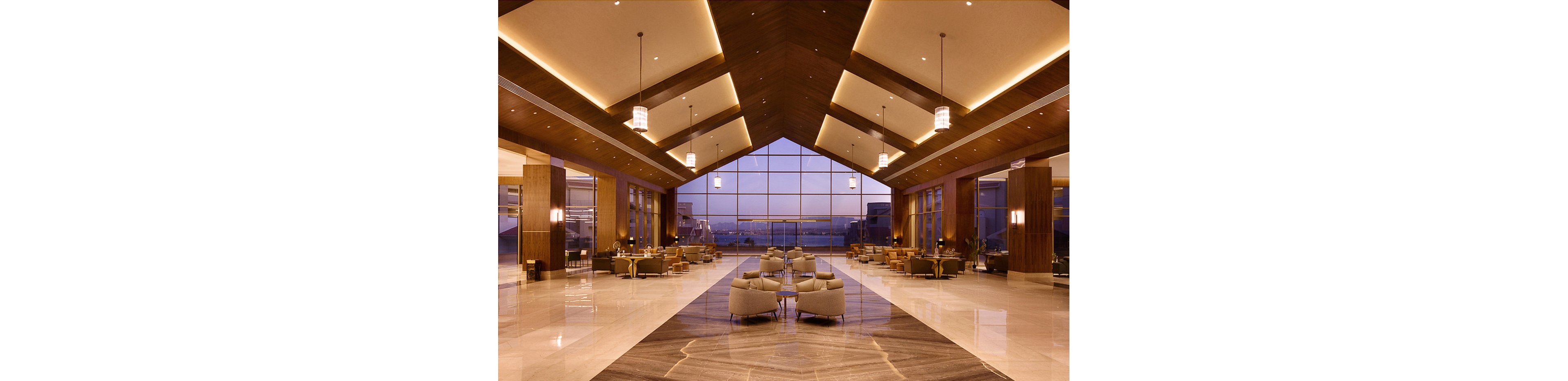 A luxurious hotel lobby features high vaulted ceilings with warm recessed lighting and elegant pendant lamps. The space is furnished with contemporary seating areas and polished marble floors, leading to a large glass wall overlooking a cityscape at dusk. The ambiance is calm and inviting, with a neutral color palette and sophisticated architectural details.