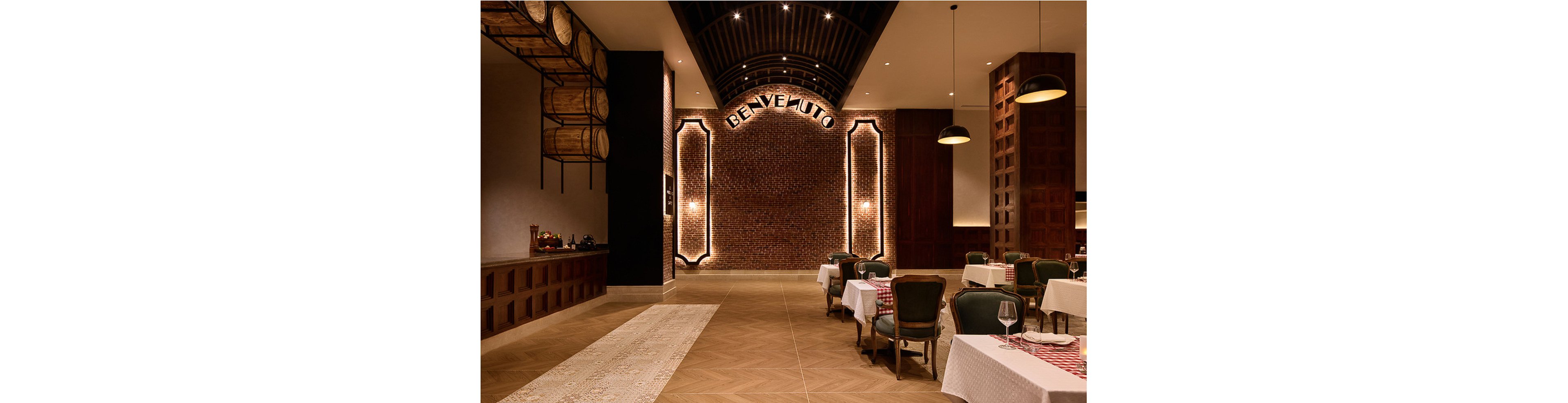 Spacious restaurant interior featuring a warmly lit brick wall with the word 'BENVENUTO' prominently displayed. The setting includes classic dining tables with white and red checkered tablecloths, vintage-style chairs, and wine barrels mounted on the wall. The ambiance is cozy and inviting, with soft lighting and a sophisticated palette of browns and neutrals.