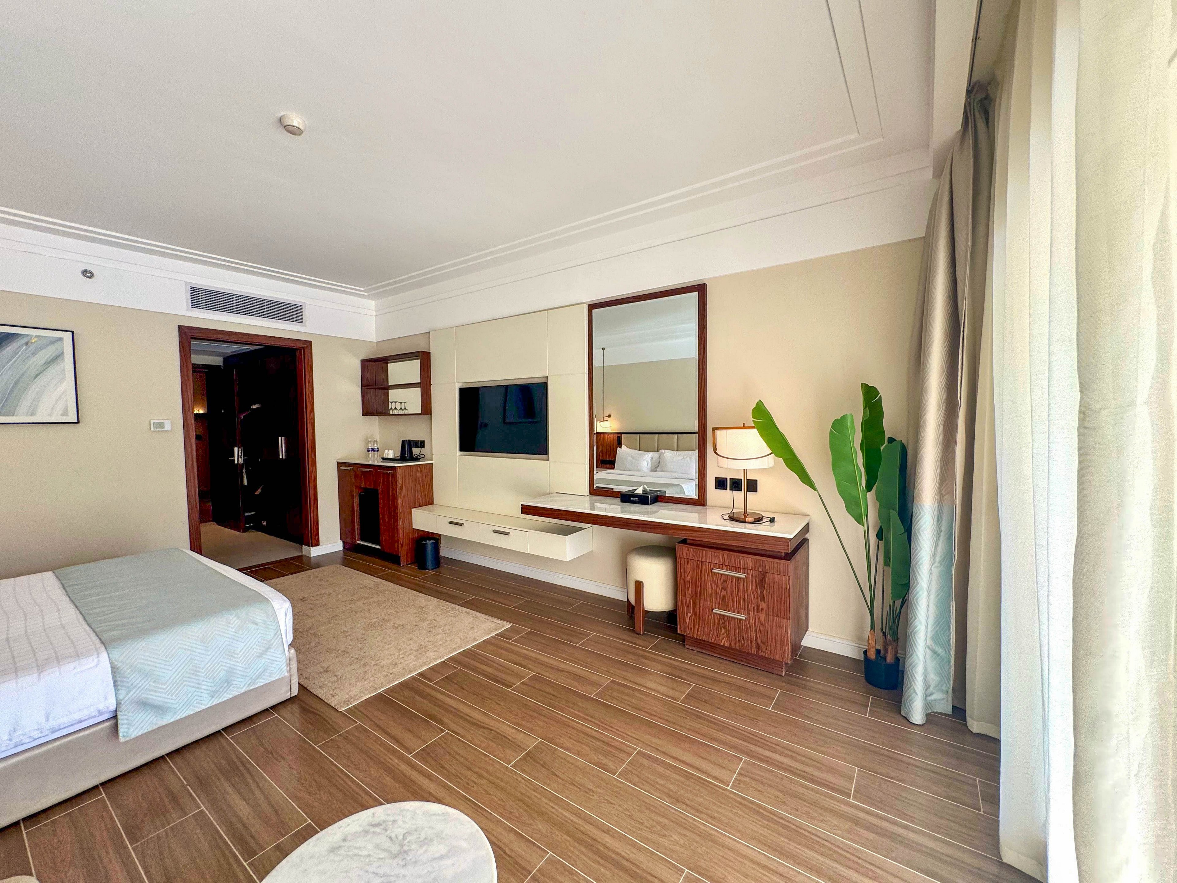 A spacious hotel room features a neatly made bed, a large wall-mounted television, and a wooden desk with a mirror. The setting is bright with natural light streaming through large curtains, and a tall green plant adds a touch of freshness. The room has a minimalist, contemporary style with neutral tones and clean lines.