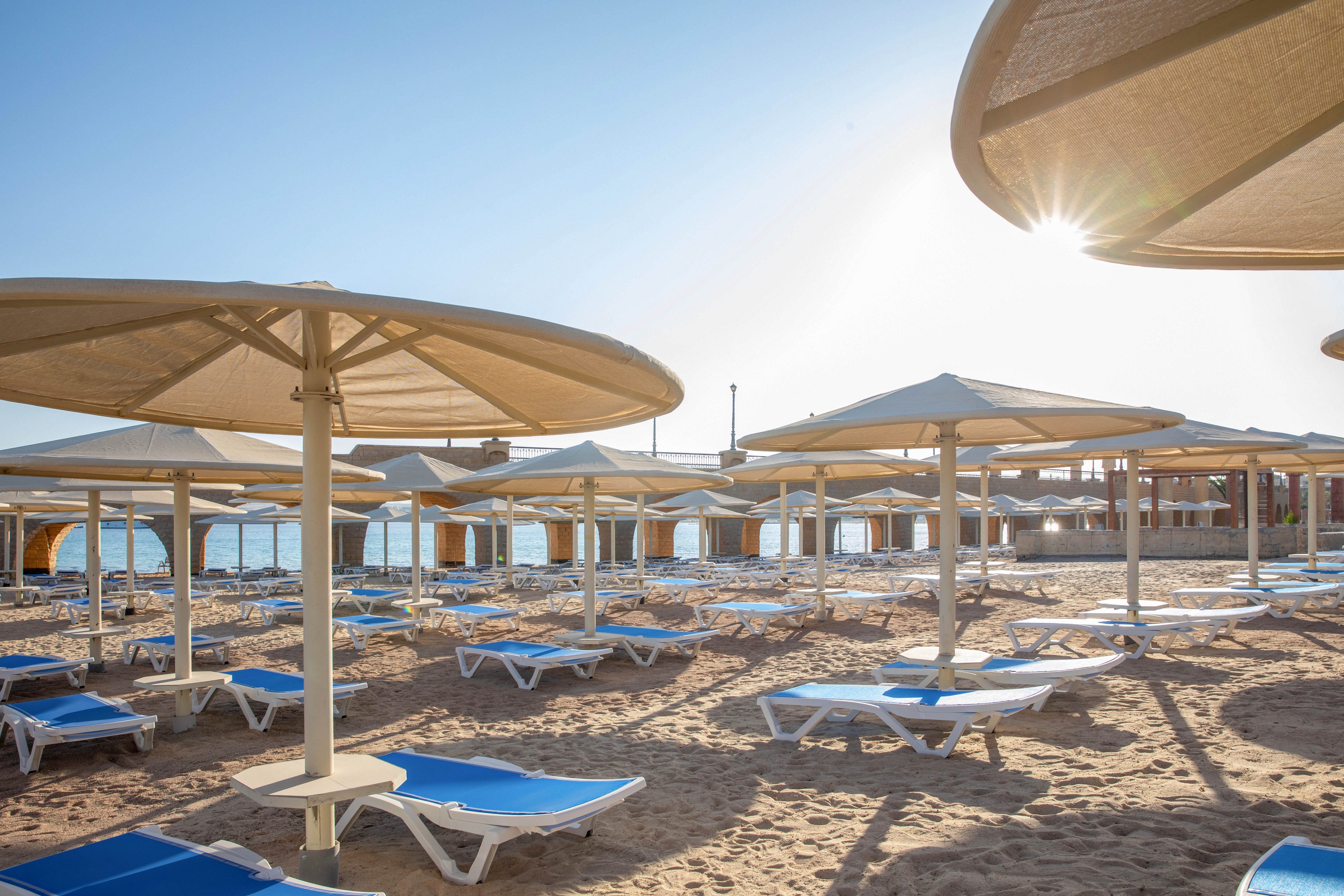 A row of blue and white beach loungers sits empty under large sun umbrellas on a sandy beach. The early morning sunlight casts long shadows, creating a tranquil and inviting atmosphere. The calm sea and a wooden pier are visible in the background, enhancing the peaceful vacation setting. No people are present, emphasizing the serene mood.
