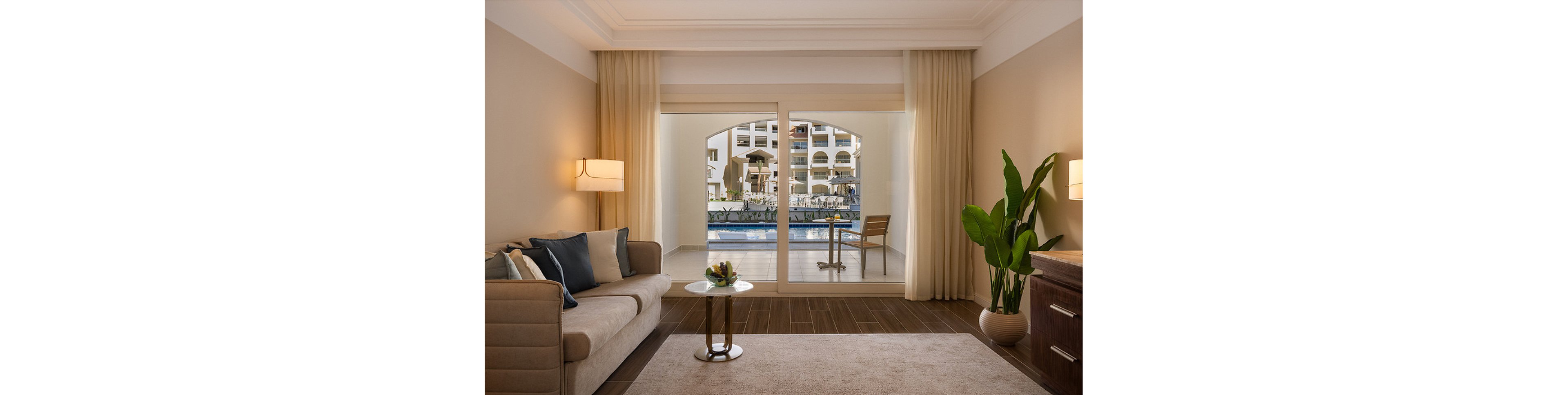 A contemporary hotel suite features a cozy seating area with a beige sofa, accent pillows, and a small round table. Large sliding glass doors open to a private terrace with a table and chair, overlooking a pool and resort buildings. The room is decorated in neutral tones with natural light, a potted plant, and elegant lighting fixtures.