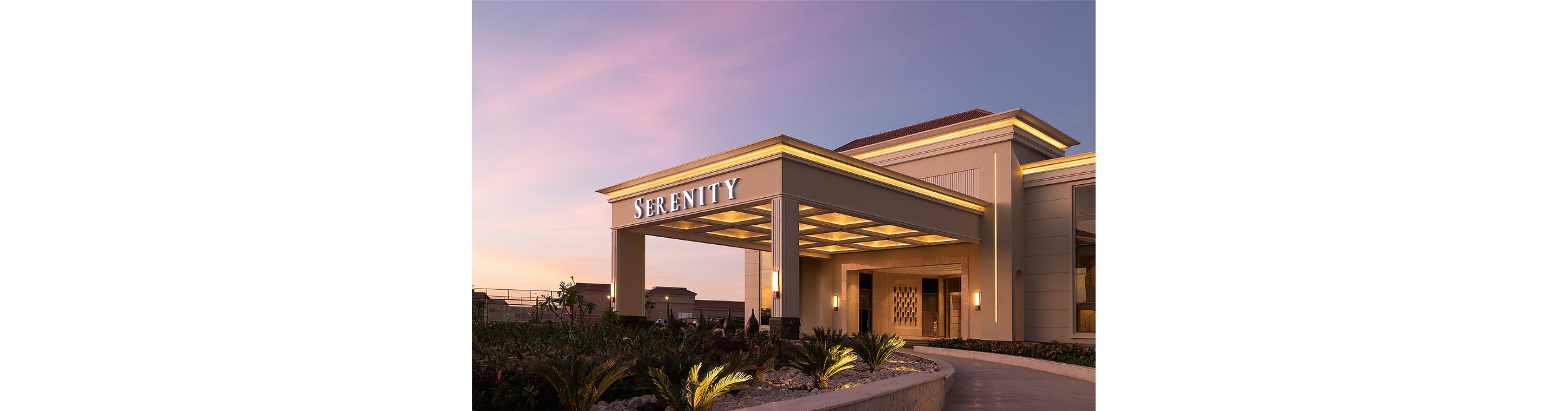 The image features the exterior of a building with the word 'SERENITY' prominently displayed in illuminated letters above the entrance. The setting is during sunset, casting a warm glow over the modern architectural structure. The foreground includes landscaped greenery, while the building's lighting enhances its inviting and tranquil atmosphere.