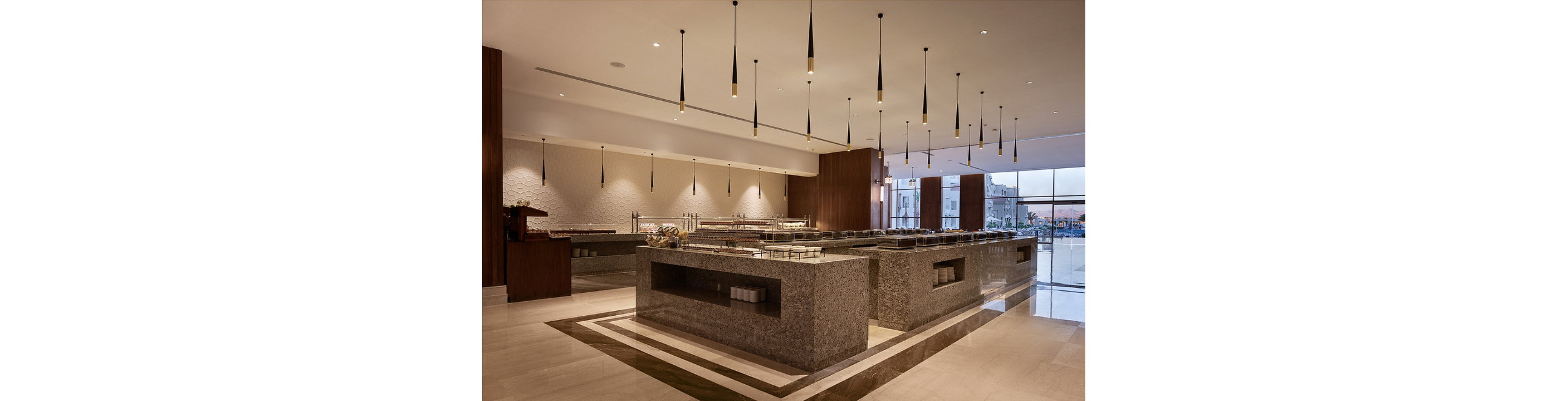 Spacious buffet area featuring sleek granite counters and contemporary pendant lighting. The setting is a modern hotel or restaurant dining space, with empty serving stations ready for food service. Warm neutral tones and clean lines create a sophisticated, inviting atmosphere. Large windows provide natural light and a view of the exterior.