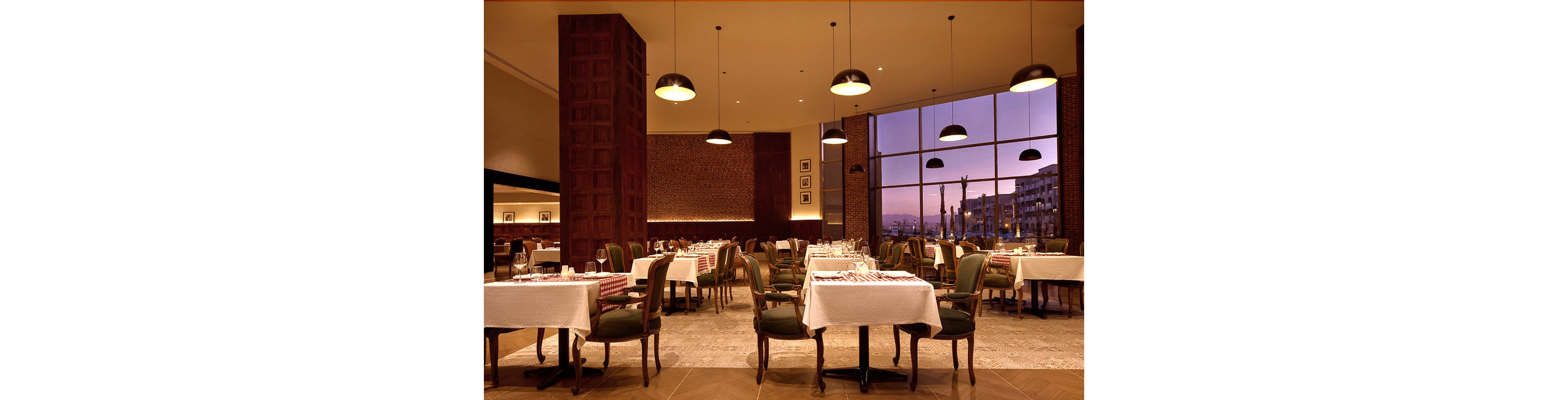 A spacious restaurant dining area features neatly arranged tables with white tablecloths and checkered runners. The setting is warmly lit with pendant lights and large windows reveal a cityscape at sunset. The atmosphere is inviting and sophisticated, with classic wooden furniture and a calm, ambient mood.